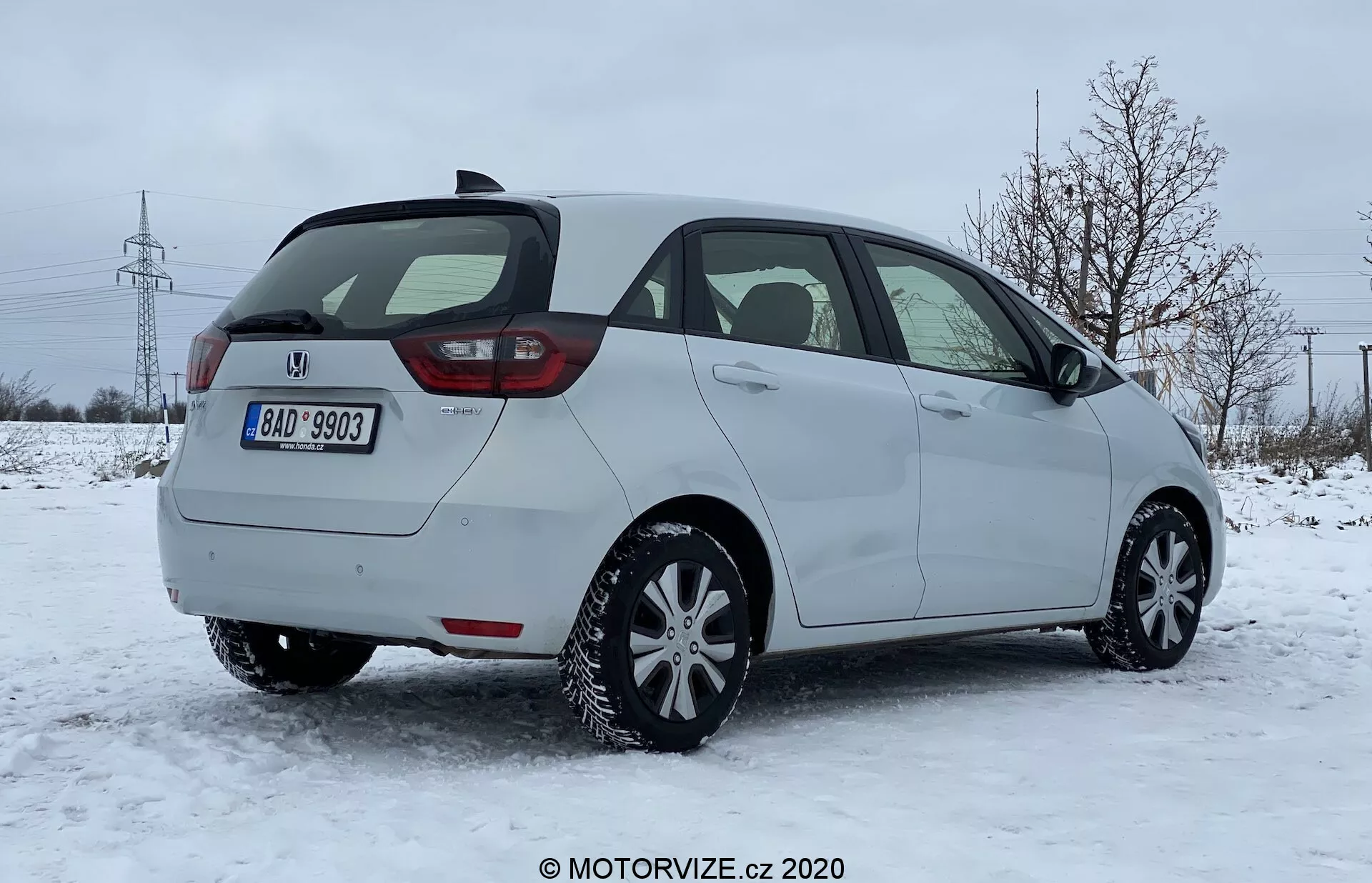 Una vista posteriore a tre quarti di un'auto hatchback Honda Jazz 2020 su una superficie coperta di neve in un ambiente suburbano con alberi spogli e torri elettriche sullo sfondo. L'immagine cattura il lato passeggero dell'auto, il design moderno dei fanali posteriori probabilmente con tecnologia LED, il portellone posteriore, il tergicristallo posteriore, il paraurti posteriore visibile con sensori di parcheggio integrati e probabilmente pneumatici invernali. Il veicolo è parcheggiato alla luce naturale con un cielo coperto, suggerendo condizioni di guida invernale.
