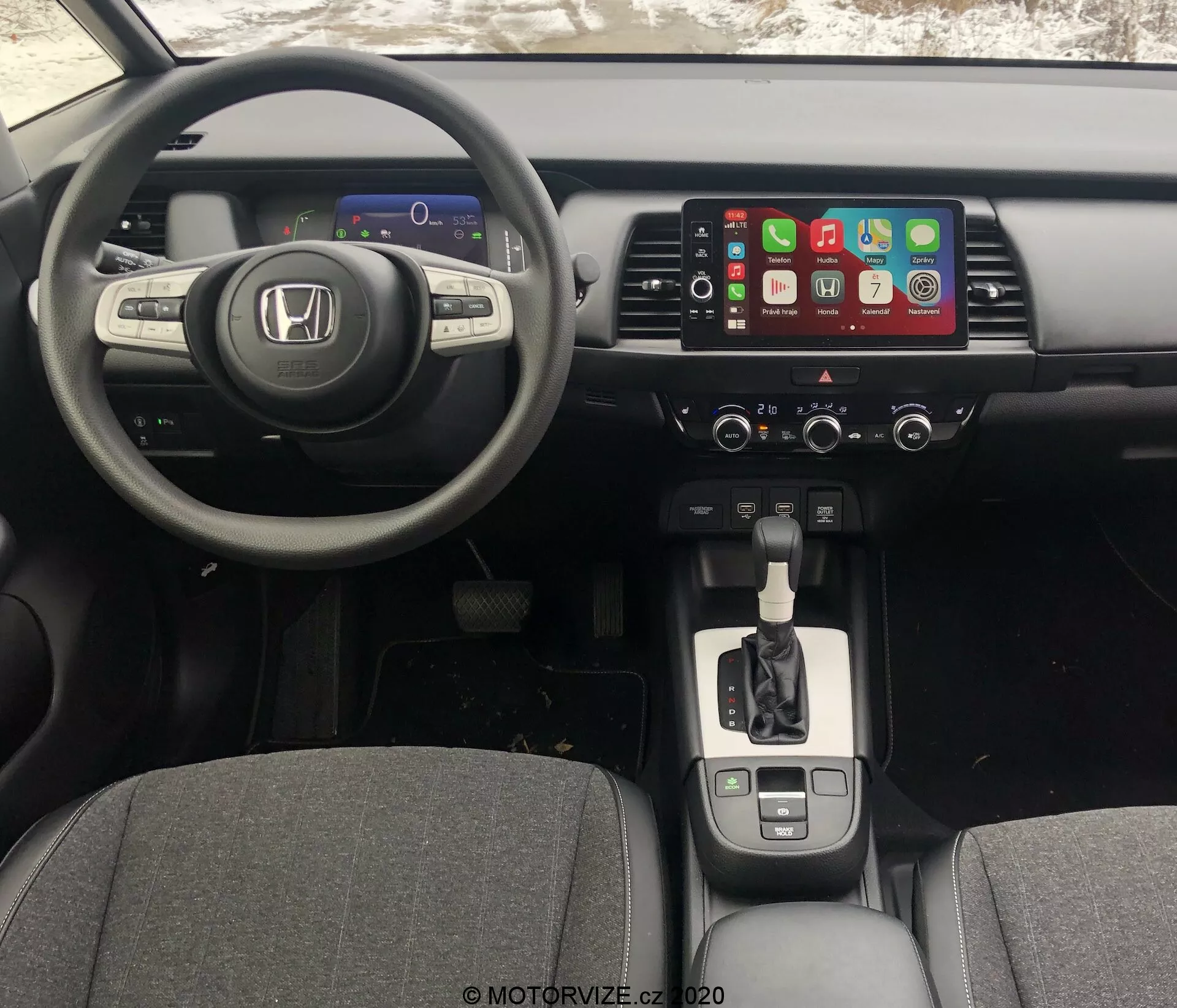 Vista interna della Honda Jazz 2020 dalla prospettiva del conducente, che mostra il cruscotto, il volante e la console centrale. L'angolo leggermente rialzato rivela il cluster digitale degli strumenti, il volante multifunzionale a tre razze, il sistema di infotainment con touchscreen compatibile con Apple CarPlay, i controlli del clima, la leva del cambio con finitura cromata, i portabicchieri, il freno di stazionamento elettronico e la connettività USB.