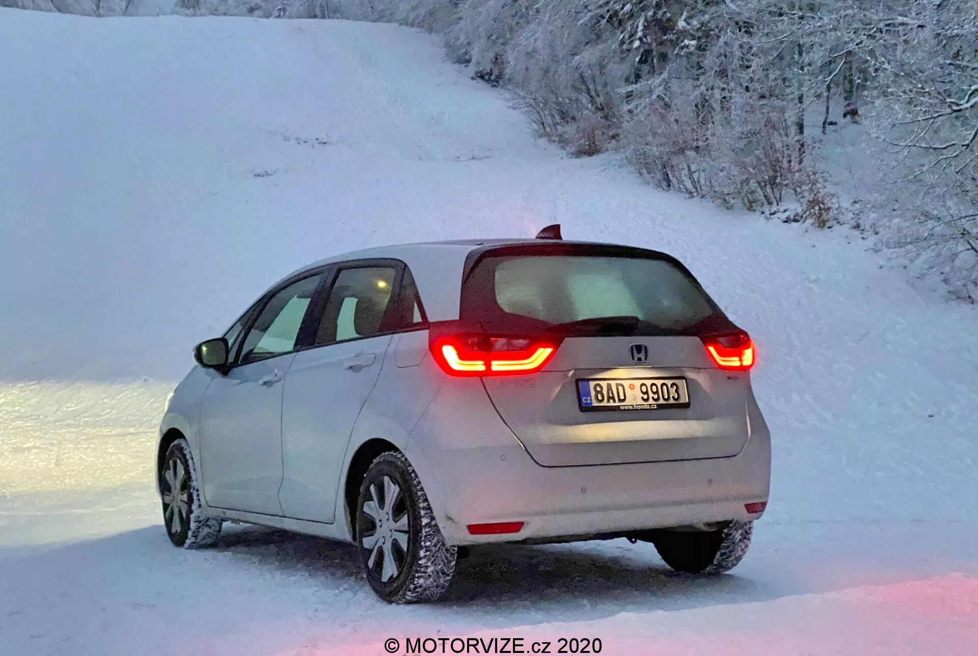 Vista posteriore tre quarti di una moderna Honda Jazz 2020 hatchback, illuminata da una soffusa luce dell'alba o del tramonto, con luci posteriori a LED illuminate di forma a C, un logo del produttore visibile, una leggera spolverata di neve sul veicolo e cerchi in lega con pneumatici coperti di neve in un ambiente rurale innevato.
