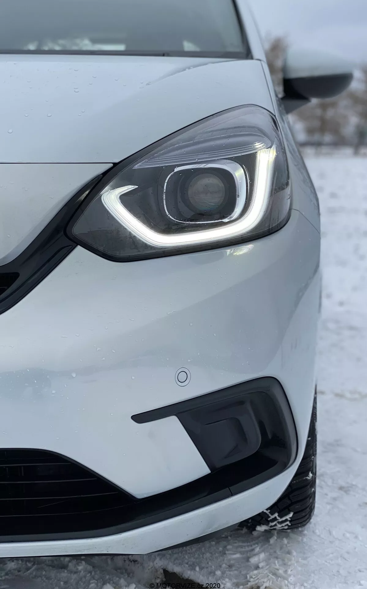 Immagine ravvicinata del lato anteriore sinistro di una Honda Jazz 2020, scattata da una bassa angolazione. Il fuoco è sul faro che presenta un design moderno con luci diurne a LED che curvano intorno alla parte superiore e laterale. L'assemblaggio del faro include una lente del proiettore visibile, suggerendo fari avanzati HID o LED per il fascio principale. Il paraurti anteriore, mostrato sotto il faro, include una griglia di presa d'aria e un alloggiamento per i fendinebbia. La vernice argento metallico o grigio chiaro della macchina e la ruota parzialmente visibile e lo specchietto laterale sono dettagliati. La neve e il ghiaccio a terra indicano un clima freddo, riflettendo le prestazioni dell'auto in condizioni invernali. L'immagine evidenzia anche la qualità costruttiva dell'auto, gli spazi tra i pannelli e il sensore di prossimità sul paraurti, che fa parte della tecnologia di assistenza al parcheggio.