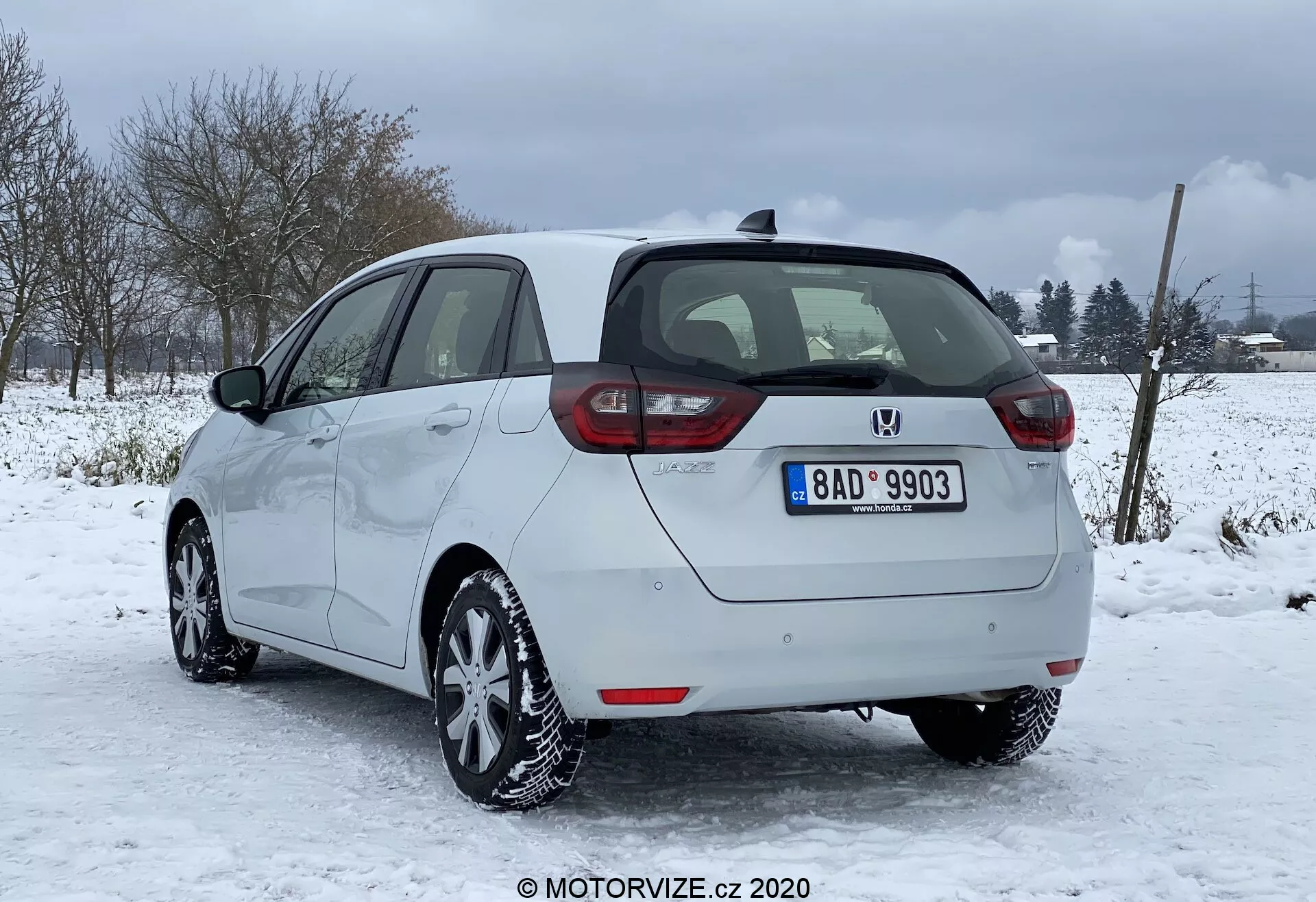 Immagine di una Honda Jazz 2020 argentata parcheggiata in condizioni nevose con un paesaggio rurale o semi-rurale sullo sfondo. Catturata dal retro e dal profilo laterale parziale destro, evidenziando i fari posteriori disposti verticalmente, il badge centrale e il paraurti con sensori di parcheggio. I contorni laterali lisci, i cerchi in lega e gli pneumatici invernali sono chiaramente visibili, insieme alla capacità di sbrinamento dei finestrini e del lunotto posteriore, e agli specchietti laterali non pieghevoli dello stesso colore della carrozzeria. Si notano la targa, l'antenna sul tetto e le buone condizioni generali del veicolo.
