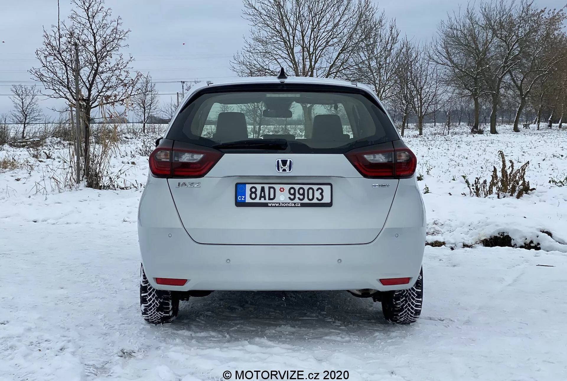 Vista posteriore di una Honda Jazz 2020 hatchback argentata parcheggiata su una superficie ghiacciata. L'immagine cattura chiaramente il paraurti posteriore, il portellone, le luci posteriori e il parabrezza posteriore del veicolo. Il logo del produttore e il nome del modello sono visibili sul portellone, insieme a una targa standard montata centralmente. L'auto presenta luci targa illuminate, riflettori su entrambe le estremità del paraurti posteriore e luci posteriori a lampadina standard. Il parabrezza posteriore ha una funzione di sbrinamento mostrata da linee orizzontali e un tergilunotto posteriore parcheggiato nella parte superiore. Il sistema di scarico è leggermente visibile sotto il paraurti posteriore sul lato destro. L'auto ha ruote in acciaio con copriruote decorative ed è ambientata in un paesaggio invernale con neve e alberi spogli sullo sfondo.