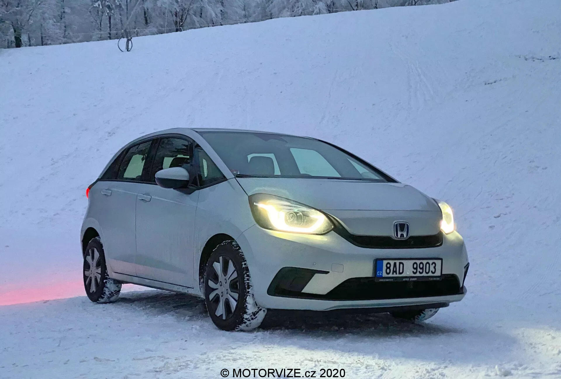 Auto hatchback Honda Jazz 2020 color argento parcheggiata su una superficie innevata all'alba o al tramonto, con fari caldi accesi, catturata da una vista tre quarti anteriore. L'immagine mette in risalto la griglia anteriore, i fari, il cofano, il parabrezza e parte del lato del conducente. Targa europea visibile, neve sul terreno e sull'auto, creando un'atmosfera fredda ma accogliente. Leggera riflessione di luce rosa sulla neve sotto l'auto. Scatto ad angolo basso che enfatizza le attrezzature per pneumatici invernali e l'aspetto dinamico per le capacità di guida in inverno.