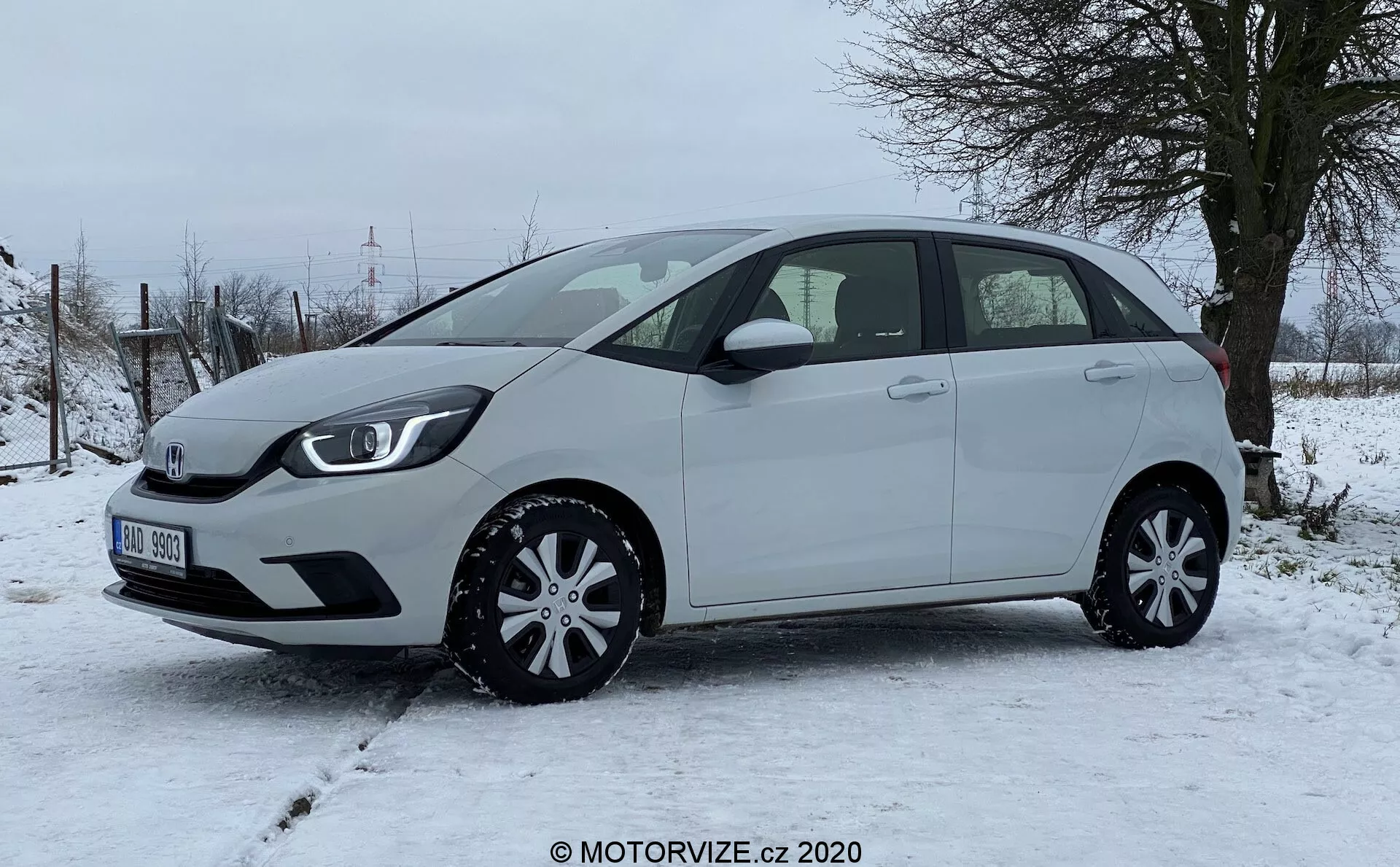 Una moderna Honda Jazz 2020 hatchback fotografata di lato, evidenziando il profilo sinistro del veicolo su un terreno nevoso con vegetazione rurale sullo sfondo. La compatta vettura presenta una linea del tetto leggermente inclinata, cinque porte inclusa la posteriore, specchietti laterali in tinta carrozzeria e copriruote eleganti, esprimendo praticità ed efficienza nel design. Nessuna caratteristica esterna avanzata come telecamere o sensori è visibile da questo angolo, sottolineando una vista chiara delle proporzioni dell'auto, contorni delle porte, linea dei finestrini e archi delle ruote.