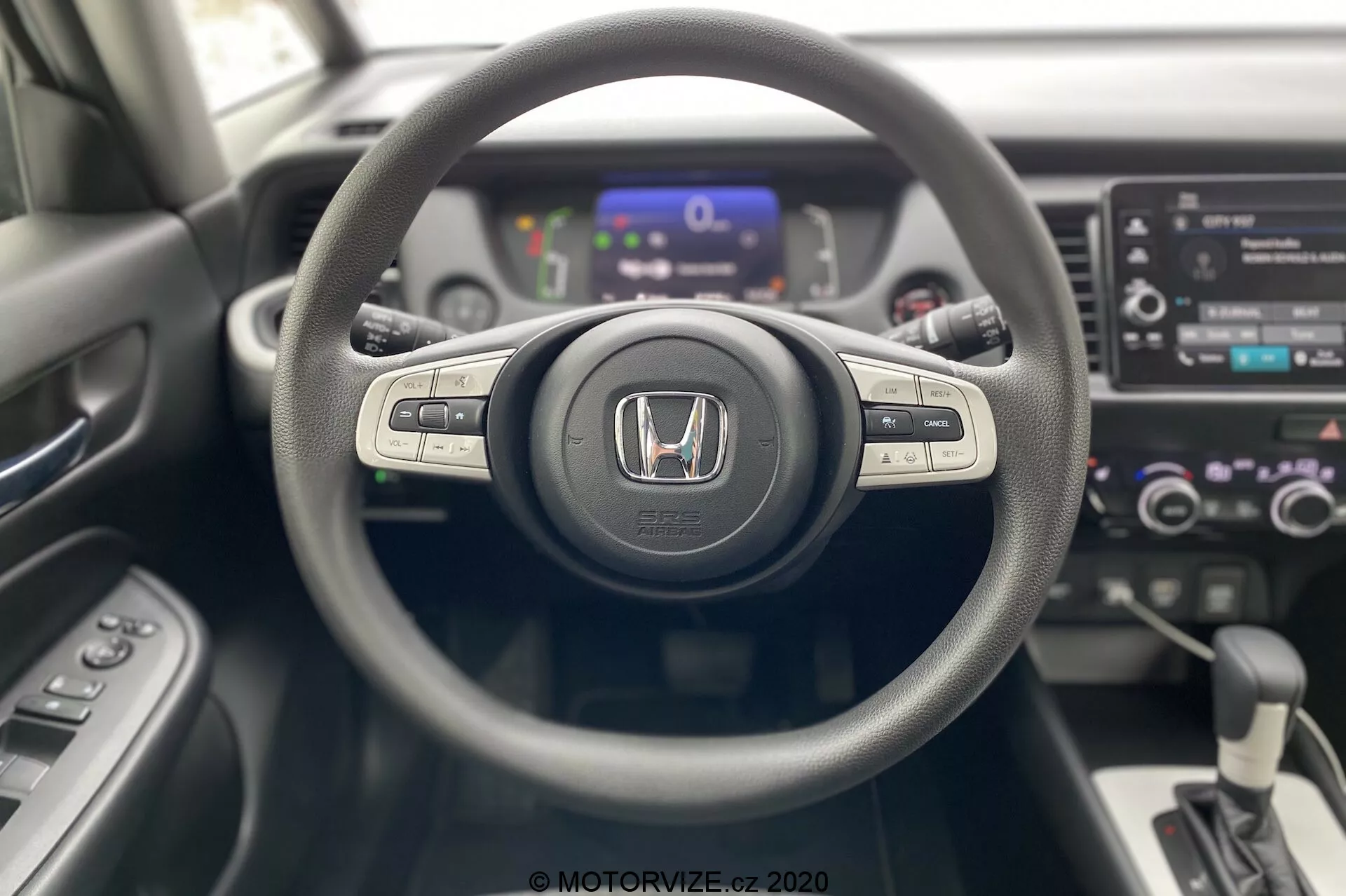 Vista interna della Honda Jazz 2020 dalla prospettiva del conducente, evidenziando il volante a tre razze con emblema Honda, pulsanti di controllo per il sistema multimediale e il cruise control sui raggi, un pannello strumenti digitale sullo sfondo leggermente sfocato e una console centrale con sistema di infotainment touchscreen, controlli del clima a doppia zona e il cambio automatico. Design moderno e contemporaneo con materiali a texture miste.