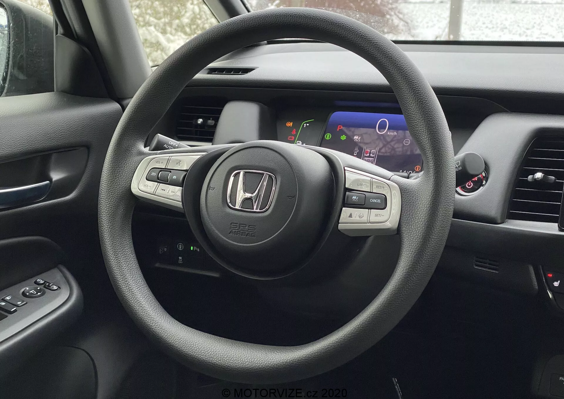 Vista interna di una Honda Jazz 2020 dalla prospettiva del sedile del conducente, evidenziando il volante a tre razze con il logo Honda e i comandi montati per audio, controllo di crociera e probabilmente Bluetooth o controllo vocale. Dietro il volante c'è un cruscotto digitale con display colorati che includono tachimetro e contagiri. Parzialmente visibile è la console centrale con schermo di infotainment o navigazione e le bocchette di controllo del clima. È anche parzialmente visibile il pannello della porta del conducente con pulsanti di controllo finestrini e specchietti laterali.