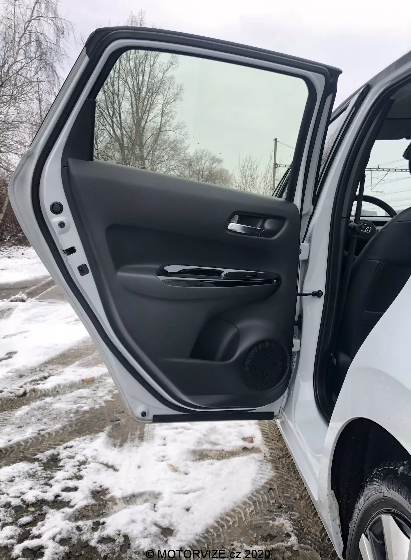 Primo piano dell'interno della porta anteriore lato guida della Honda Jazz 2020, che mostra una vista dettagliata della rifinitura in plastica testurizzata, maniglia ergonomica satinata, comandi dei finestrini e specchi elettrici, tasca portaoggetti modellata e griglia dell'altoparlante, il tutto in un ambiente invernale innevato.