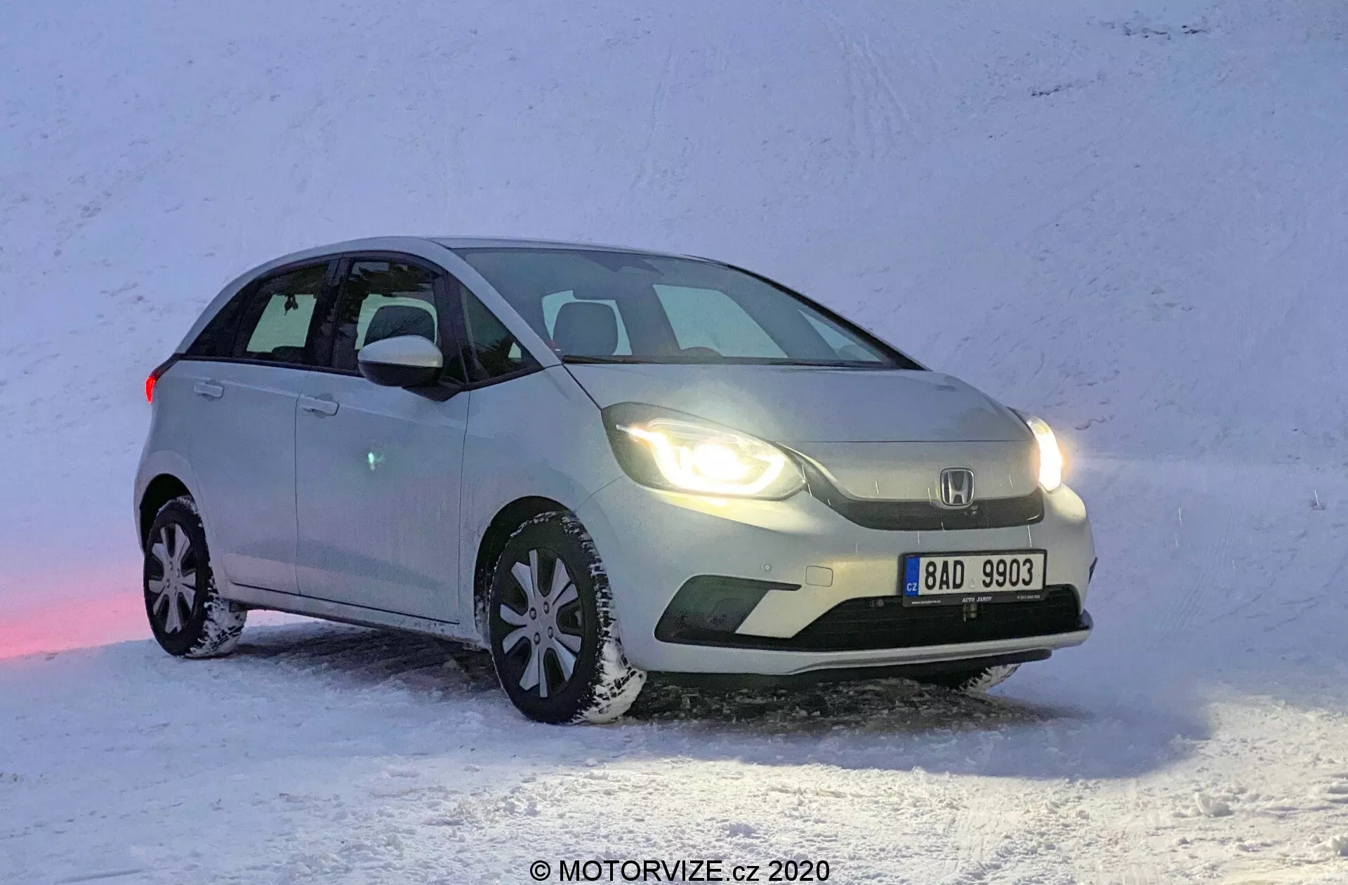 Vista frontale tre quarti di una Honda Jazz 2020 chiara, posizionata su una superficie nevosa. L'immagine cattura la macchina dal lato anteriore sinistro, mostrando il lato del guidatore ad un angolo che evidenzia sia il profilo laterale che la parte anteriore. La Honda Jazz è parcheggiata con il frontale leggermente rivolto verso destra, rivelando il design della griglia, i fari e parte del cofano. I fari anteriori sono accesi, riflettendosi sulla neve, con il veicolo apparentemente fermo. I pneumatici da neve con distinti disegni del battistrada sono visibili, sottolineando la sua idoneità per la guida invernale.