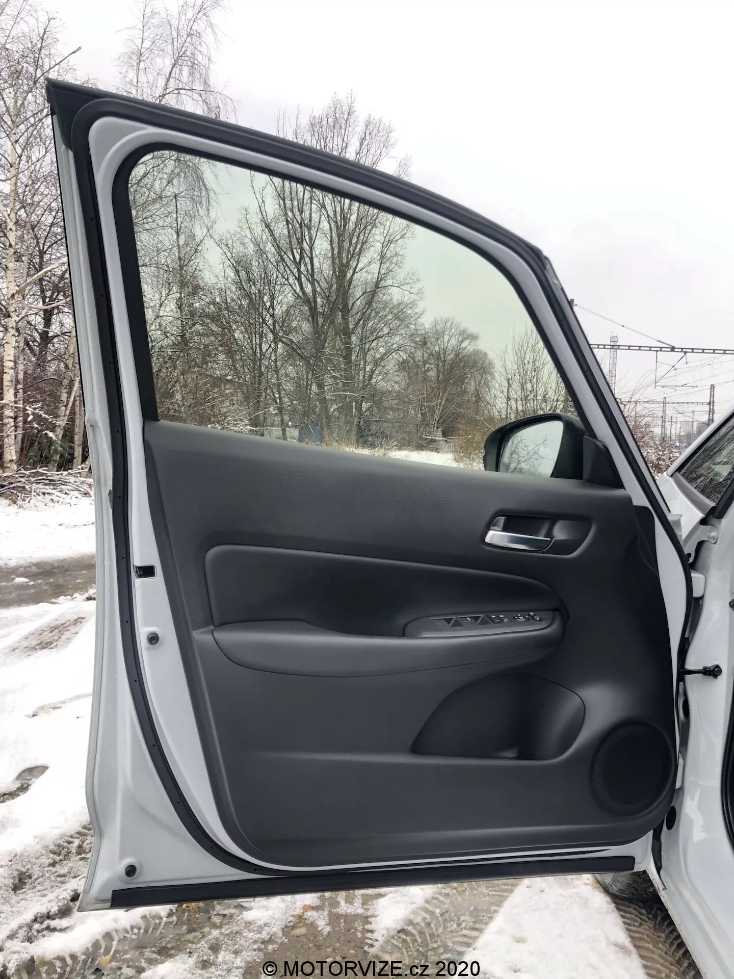 Vista interna della porta anteriore del passeggero della Honda Jazz 2020, completamente aperta, con materiali plastici neri sia testurizzati che lisci. Il pannello della porta include un bracciolo ergonomico, una maniglia morbida e un pannello di controllo per i meccanismi elettrici di finestra e serratura. È presente anche un accento trim argentato e una tasca di stoccaggio inferiore. Attraverso la finestra si vede un paesaggio innevato, che indica un ambiente invernale.