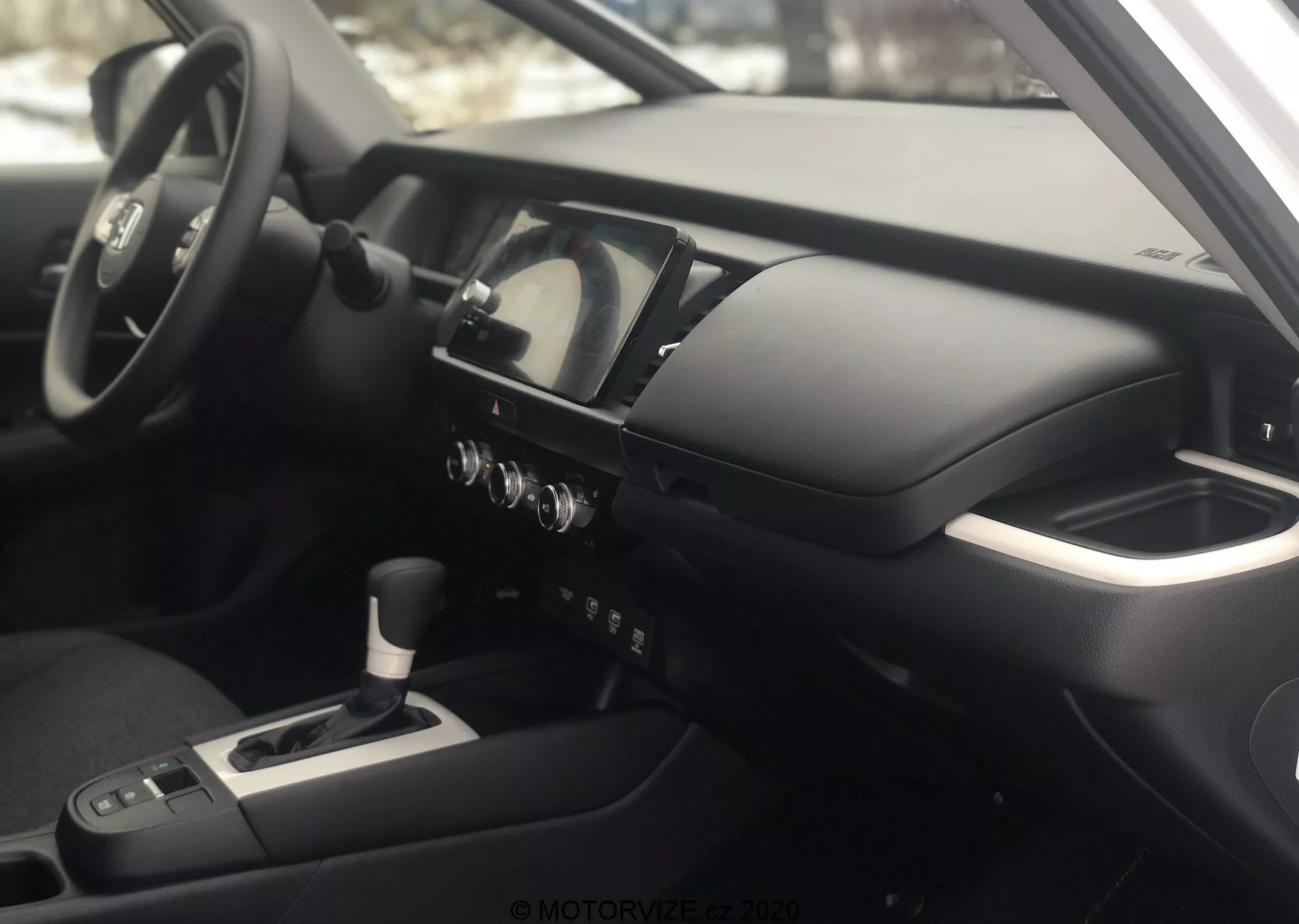 Vista interna della Honda Jazz 2020 dal lato passeggero, con attenzione al cruscotto e all'area della cabina del conducente. La foto, scattata con un angolo leggermente elevato, mette in risalto la consolle centrale, il volante multifunzione con comandi montati, il quadro strumenti digitale-analogico, la consolle centrale con cambio, i pulsanti di controllo del clima e un display di infotainment/navigazione. Il bracciolo suggerisce il comfort del conducente e l'interno è caratterizzato da materiali metallici e soft-touch, riflettendo una filosofia di design moderna e di alta gamma.