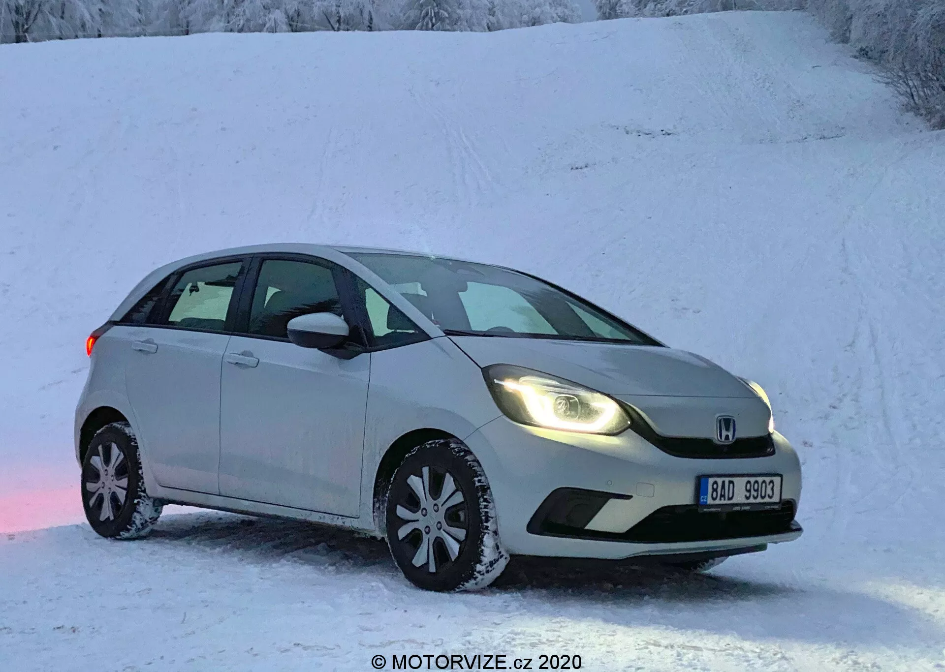 L'immagine mostra una Honda Jazz 2020, una berlina argentata o grigio chiaro parcheggiata su una superficie innevata, suggerendo una scena invernale con un paesaggio coperto di neve. Catturato da un angolo frontale a tre quarti, la vista mette in evidenza il lato del guidatore, mostrando la griglia anteriore, i fari, il cofano, il parabrezza, la porta del conducente e la porta posteriore del passeggero. L'auto presenta fari moderni, un paraurti anteriore scolpito, un badge prominente sulla griglia, specchietti laterali con indicatori di direzione integrati e cerchi in lega multispettrata dotati di pneumatici invernali. Il veicolo stazionante è raffigurato senza sfocatura del movimento in condizioni di luce bassina, o all'inizio della mattina o nel tardo pomeriggio.