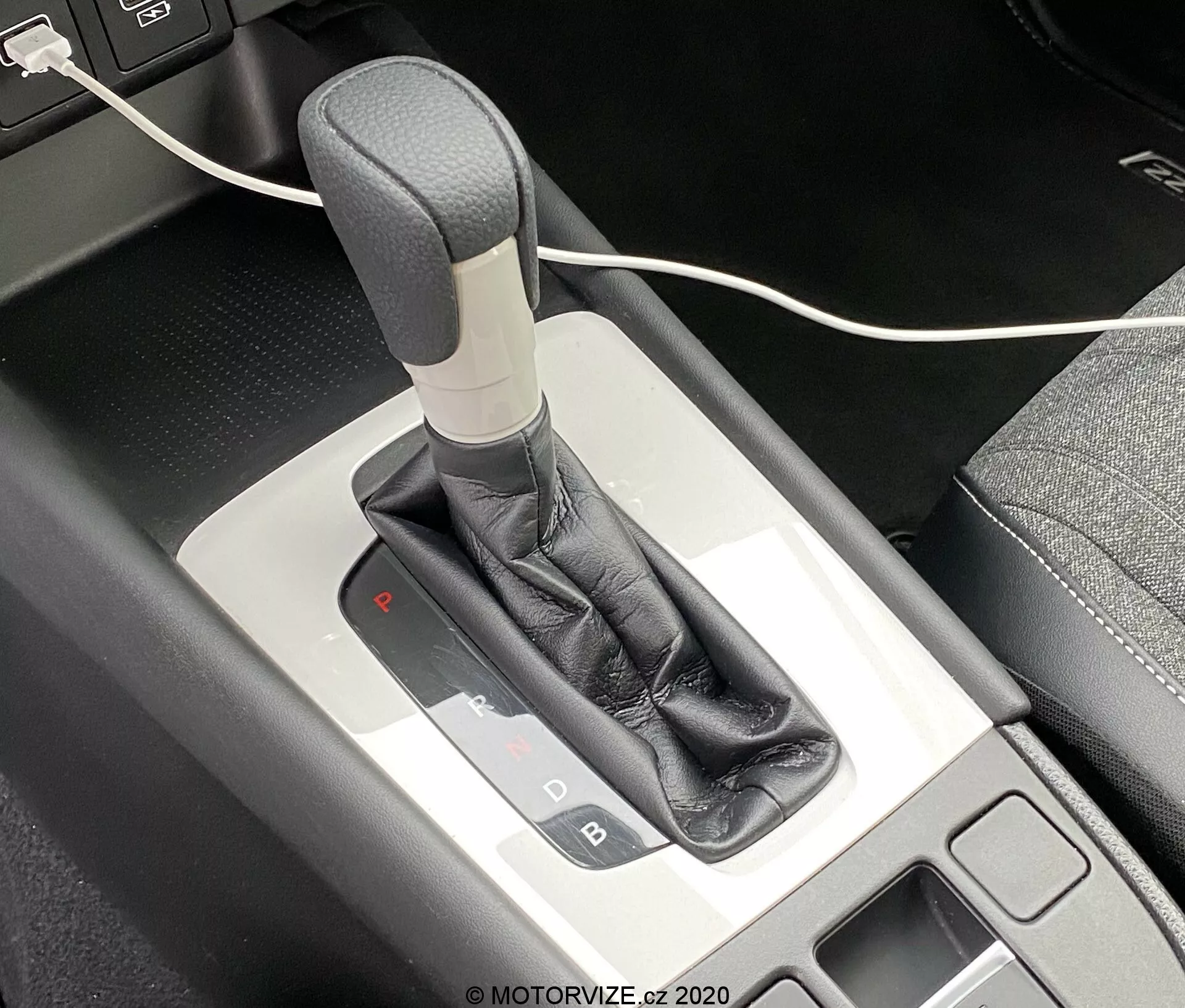 Vista ravvicinata della leva del cambio automatico della Honda Jazz 2020 da un'angolazione dall'alto, che mostra il meccanismo del selettore del cambio e la console centrale circostante. La leva del cambio presenta un manico grigio o argento, avvolto in un rivestimento in pelle nera e completato da una finitura metallica. Il pannello di selezione del cambio illuminato indica che il veicolo è in modalità parcheggio con una luce rossa sotto la 'P'. A sinistra è visibile un piccolo vano portaoggetti con un tappetino nero, insieme a un cavo USB parzialmente visibile nella parte superiore dell'immagine.