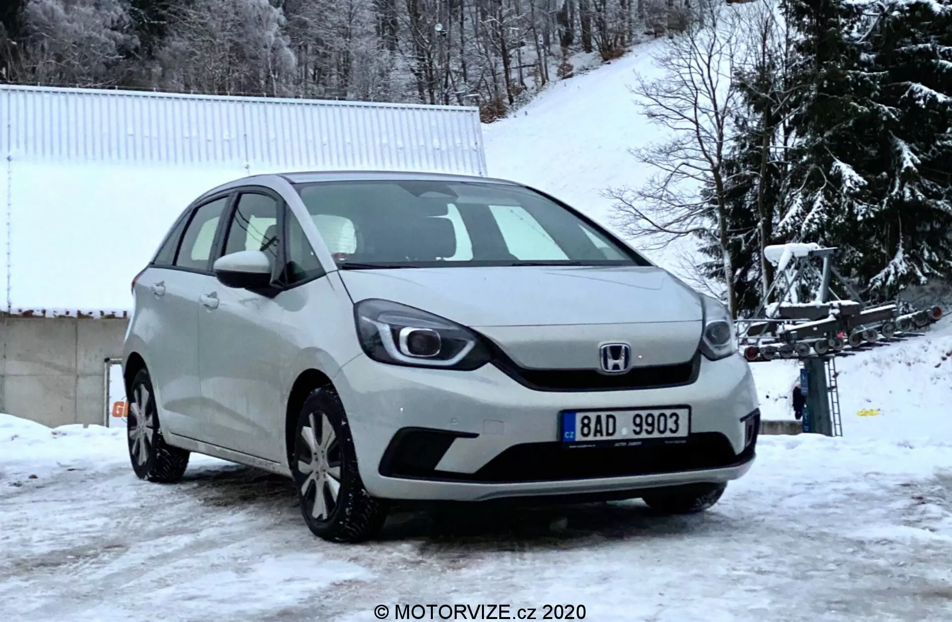 Vista di angolo frontale di una Honda Jazz argento 2020 catturata in una giornata invernale, che mostra la griglia anteriore, i fari, il cofano e il paraurti anteriore su uno sfondo innevato con alberi spogli e un edificio bianco.