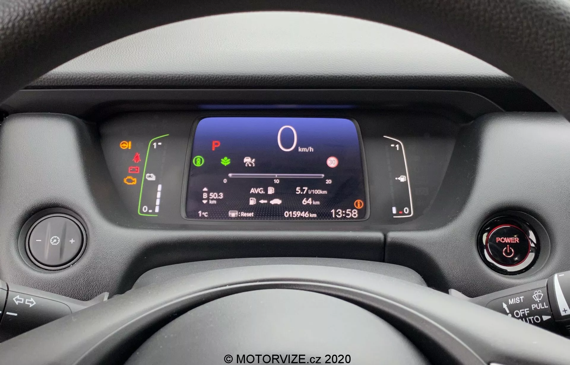 Vista dal sedile del conducente di una Honda Jazz 2020, focalizzata sul quadro strumenti moderno, volante e controlli del cruscotto. Il display digitale mostra informazioni chiave del veicolo, tra cui velocità, selezione della marcia, efficienza del carburante, ora e temperatura, con accensione senza chiave e controlli multifunzione sul volante visibili.
