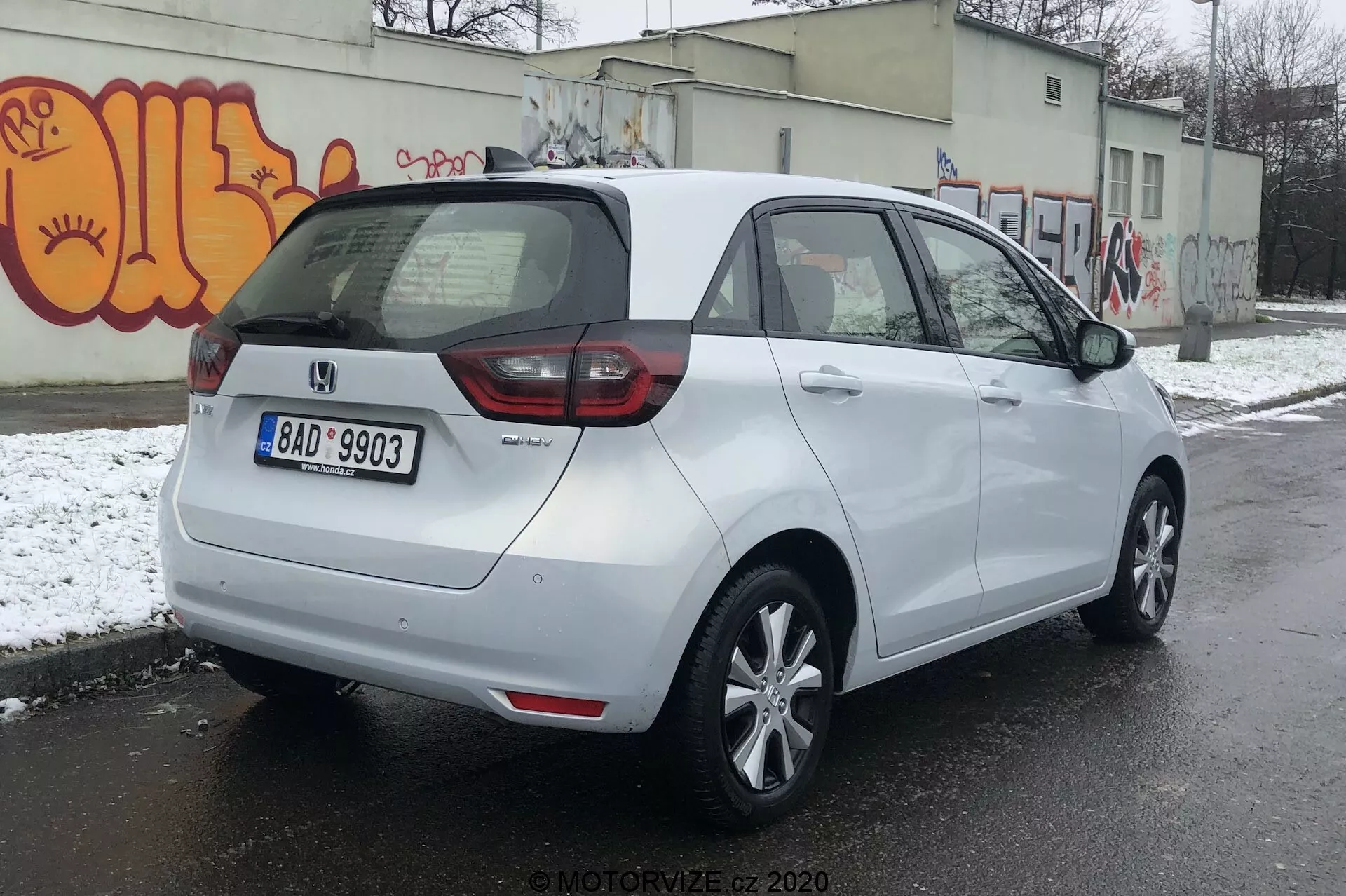 Vista posteriore di tre quarti di una Honda Jazz 2020 bianca parcheggiata su una superficie asfaltata in un contesto urbano potenzialmente freddo con neve per terra e graffiti sul muro. L'auto presenta un mix di luci posteriori a LED e tradizionali, targa posteriore centrale e un distintivo che indica il livello di allestimento o variante. Ha un tetto aerodinamico, cerchi in lega a cinque razze, specchietti laterali in tinta carrozzeria con indicatori di svolta integrati e maniglie delle porte abbinate al colore della carrozzeria. Il parabrezza posteriore è dotato di sbrinatore e tergicristallo per una migliore visibilità.