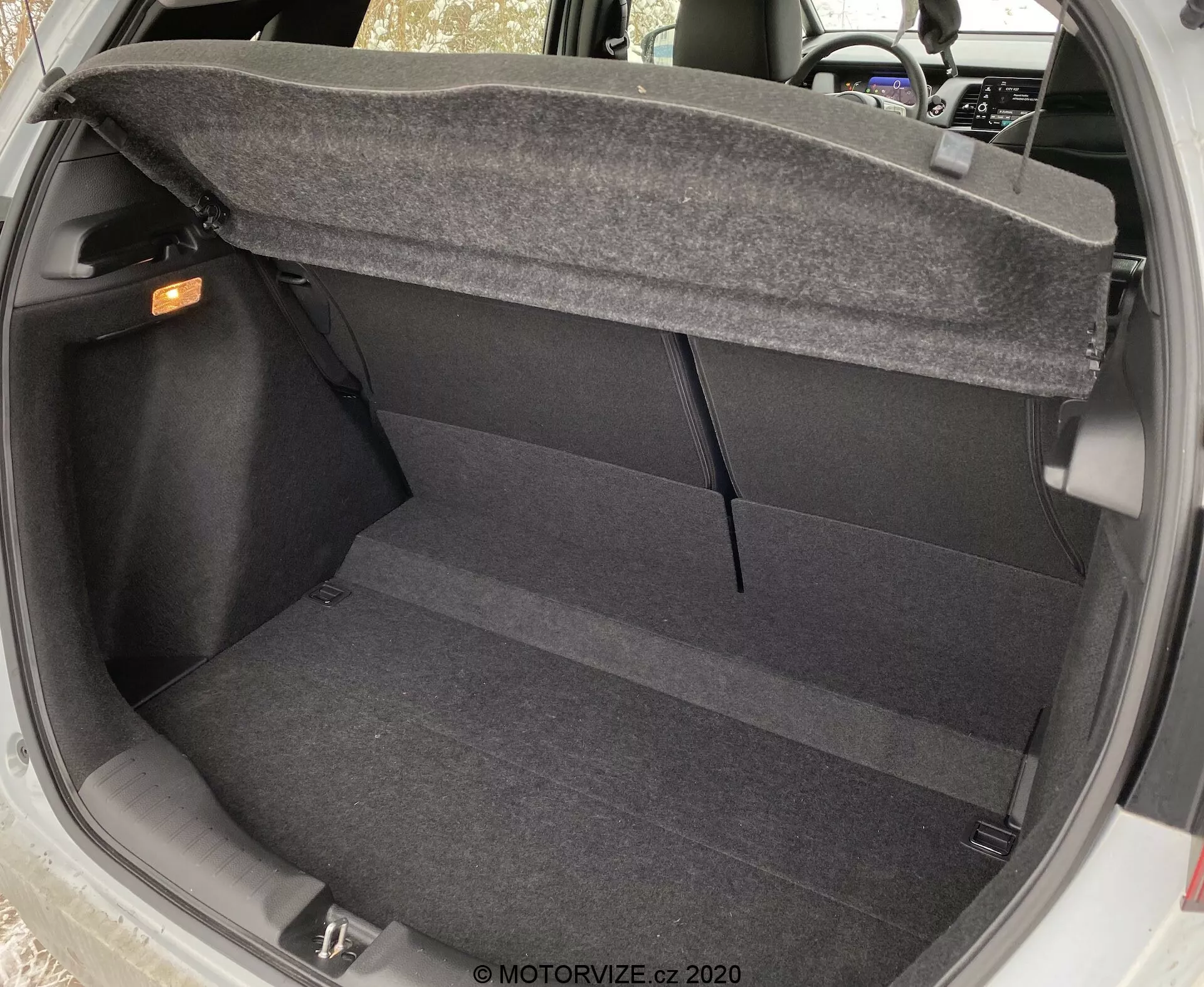 Vista interna del bagagliaio della Honda Jazz 2020 da un'angolazione leggermente elevata, che mostra il pavimento pulito e rivestito di moquette, le pareti laterali e il retro dei sedili posteriori. Il bagagliaio è vuoto, con la maniglia di rilascio interna visibile nell'angolo in alto a destra e il meccanismo di cerniera del coperchio del bagagliaio parzialmente visibile sui lati superiori, evidenziando la praticità e le caratteristiche di design di base del bagagliaio.