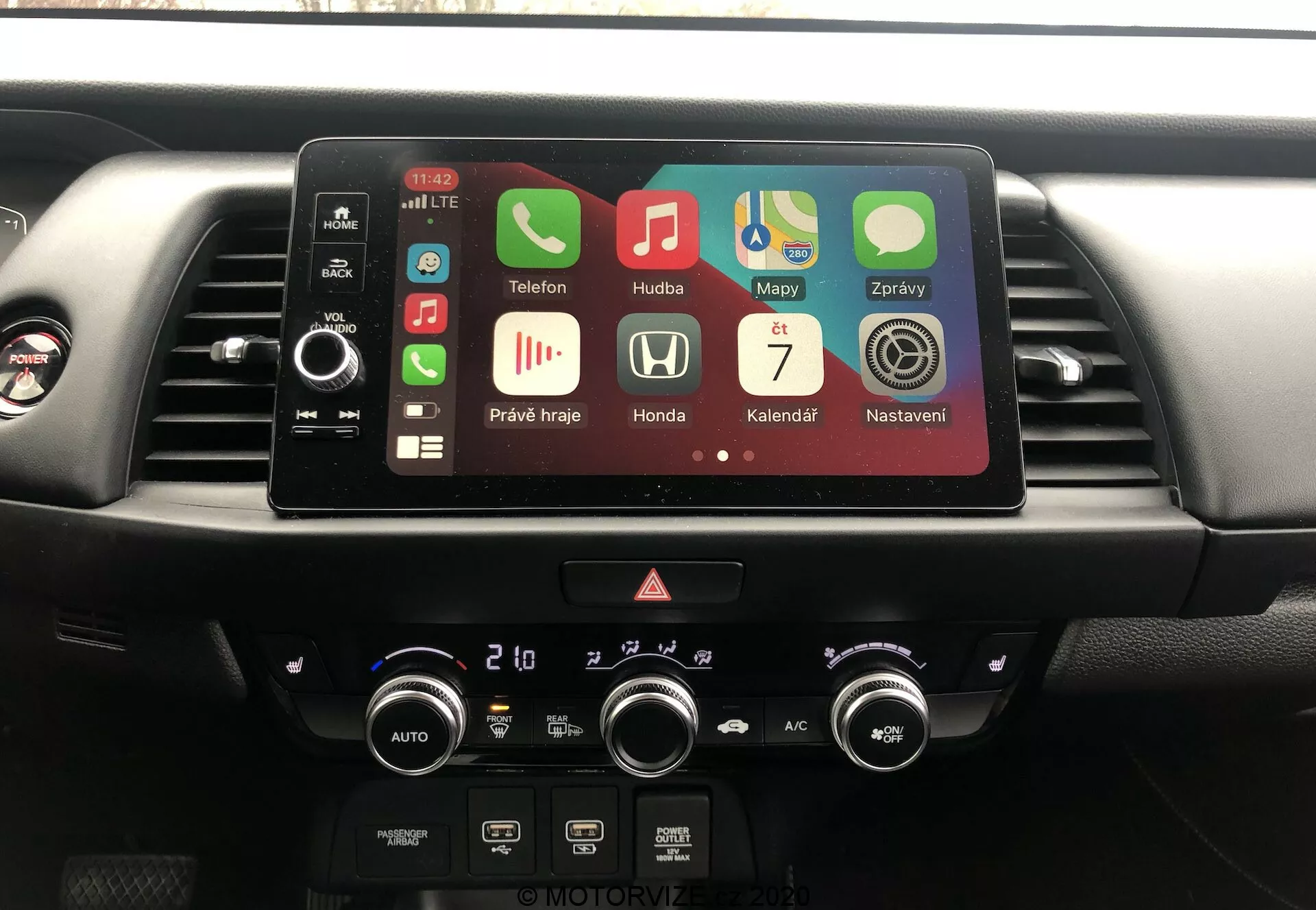 Consolle centrale dell'interno della Honda Jazz 2020, mostrando il sistema di infotainment e il controllo del clima dalla prospettiva del conducente. Display touchscreen attivo con app come Telefono, Musica, Mappe che indicano Bluetooth, GPS, riproduzione multimediale e Apple CarPlay. Sotto il display, pulsanti fisici, luci di emergenza e quadranti del climatizzatore a due zone con letture in Celsius.