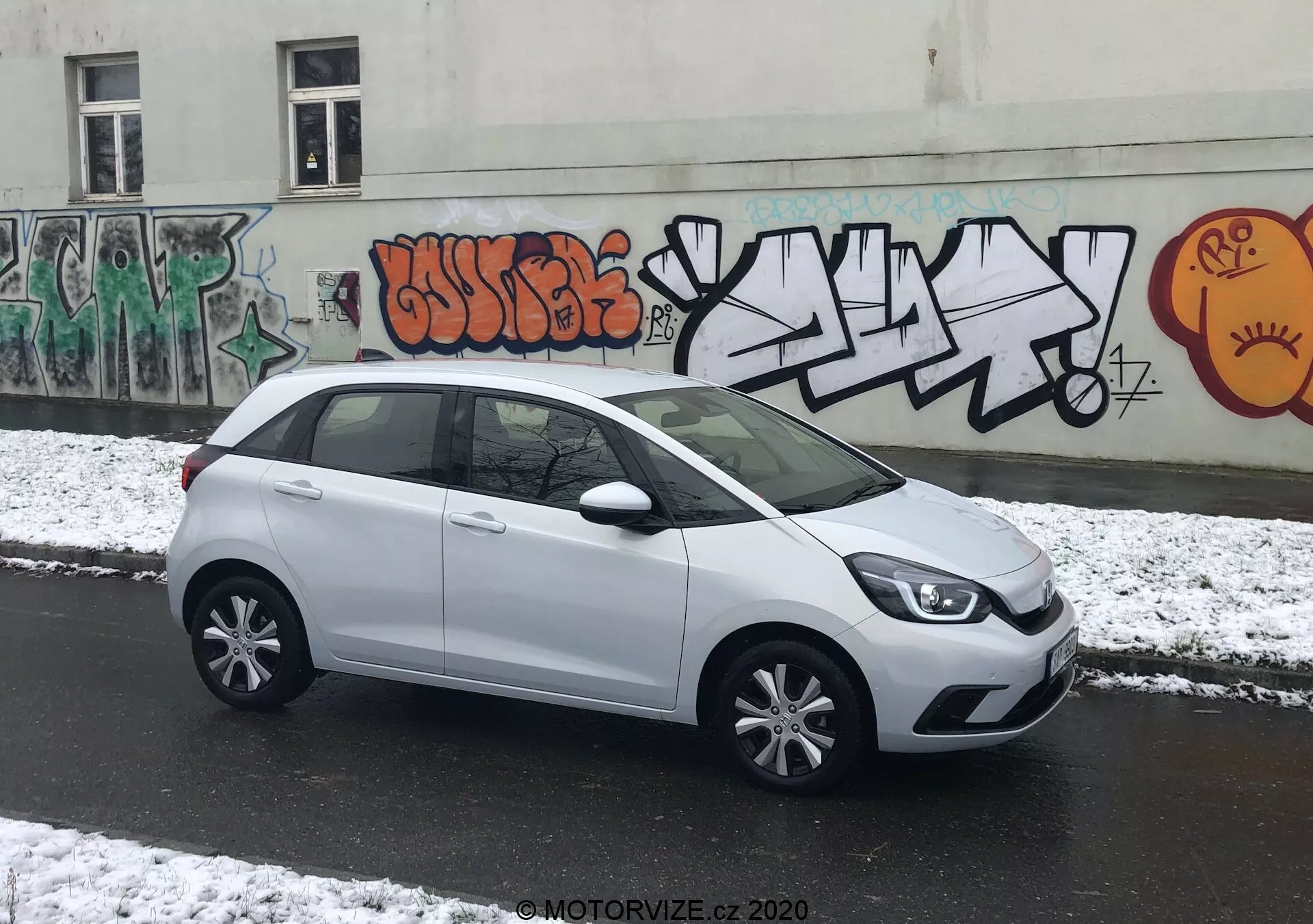 L'immagine presenta una Honda Jazz 2020 bianca parcheggiata perpendicolarmente alla fotocamera su una strada urbana, permettendo una vista laterale completa. La parte anteriore stilizzata della vettura con cluster di fari moderni è parzialmente visibile, conducendo verso una linea caratteriale morbida sul lato. Le maniglie delle porte e gli specchietti laterali in tinta con la carrozzeria e i copricerchi a più razze migliorano la sua apparenza elegante. La spaziosa finestratura offre ampio spazio per la testa, e un piccolo finestrino posteriore aggiunge al design. Lo spoiler posteriore suggerisce efficienza aerodinamica. L'illuminazione nuvolosa con neve fresca e il fondale urbano con graffiti aggiunge un contesto dinamico.