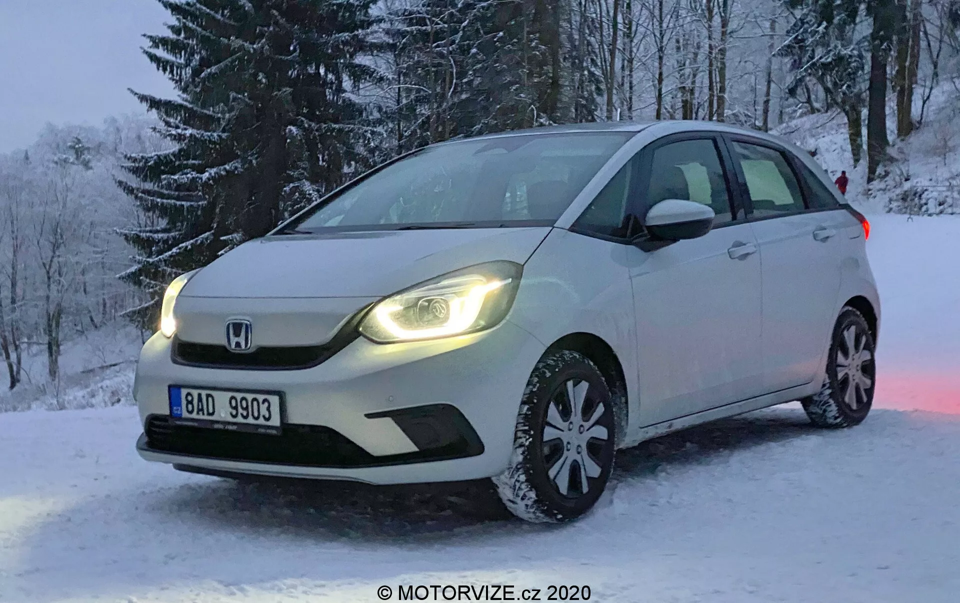 Fotografia professionale di una Honda Jazz bianca del 2020 parcheggiata su un terreno innevato durante l'inverno. L'immagine è scattata da un angolo che mette in risalto il lato anteriore sinistro dell'auto, mostrando la griglia anteriore, i fari a LED, il cofano, la ruota anteriore sinistra multi-razza con pneumatici da neve, il parabrezza e la portiera del conducente. Gli alberi innevati sono visibili sullo sfondo e la targa è leggibile. La luce soffusa suggerisce l'alba o il tramonto, con le luci di marcia diurna dell'auto che si riflettono sulla neve, migliorando l'ambiente della scena. Il veicolo è dotato di illuminazione avanzata e indicatori di direzione integrati negli specchietti laterali, con il distintivo logo Honda posizionato in modo prominente sulla griglia.
