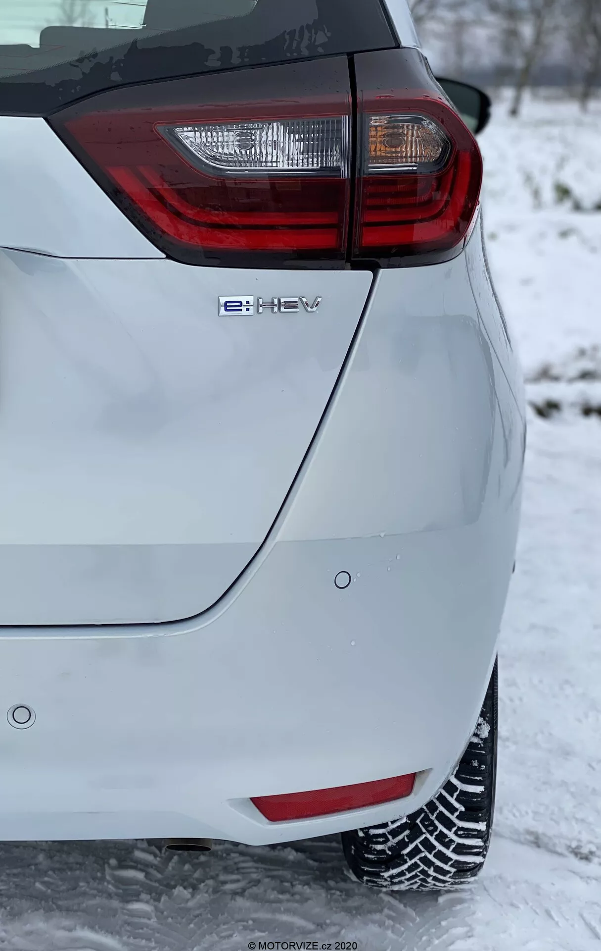 Primo piano dell'angolo posteriore di un'auto hatchback moderna, che presenta un gruppo di luci posteriori prominente con elementi rossi e chiari. L'immagine, scattata in orientamento verticale, mostra l'emblema della tecnologia ibrida 'e-HEV' ed è ambientata in un contesto invernale con neve sul terreno e che copre i pneumatici, indicando condizioni di guida invernali. La vernice di colore chiaro del veicolo contrasta con lo sfondo innevato, evidenziando le sue caratteristiche di design moderno ed ecologico.