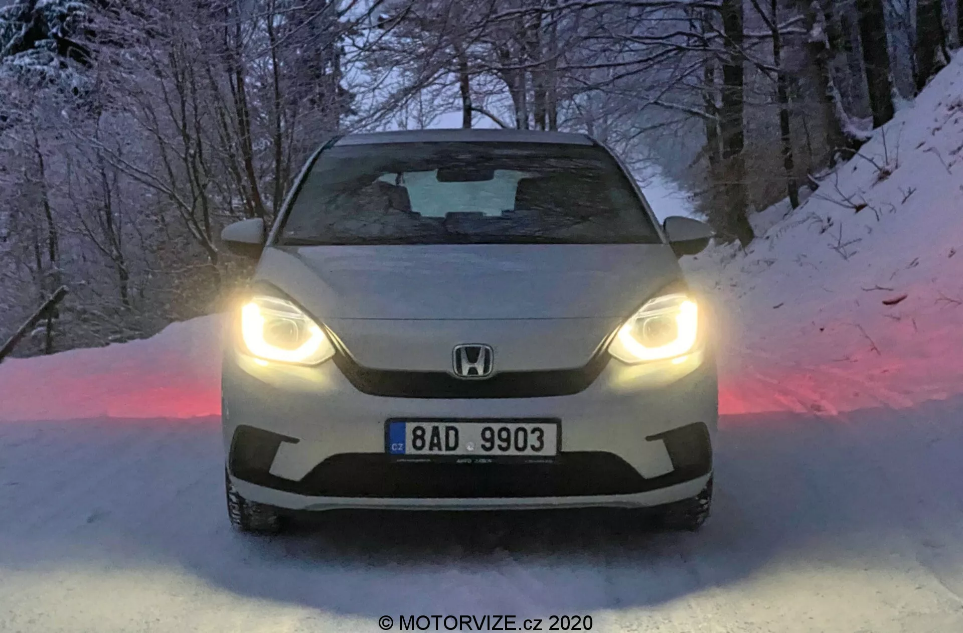 Vista frontale di una Honda Jazz 2020 illuminata con fari a LED, che mostra il logo del marchio e la targa sulla griglia, con un cofano scolpito e parabrezza oscurato. Il terreno innevato e l'atmosfera crepuscolare esaltano il design avanzato e la capacità di guida invernale dell'auto.