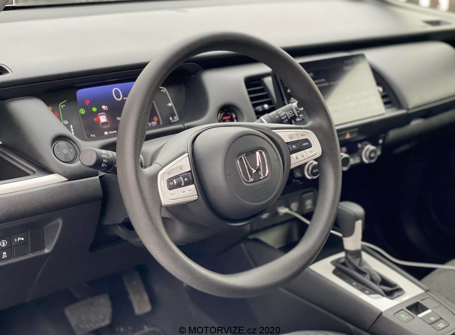 La foto mostra l'interno di una Honda Jazz 2020 dalla prospettiva del conducente. Il focus è sul volante con marchio Honda, che presenta diversi pulsanti di controllo suggerendo funzionalità integrate di audio, display informativo e possibilmente controllo di crociera. Dietro al volante, è visibile un display digitale parziale che mostra un indicatore del livello della batteria, suggerendo che il veicolo potrebbe essere ibrido o completamente elettrico. L'interno ben illuminato e moderno include anche una console centrale con un sistema di infotainment touchscreen, controlli del clima e una leva del cambio di design contemporaneo con trasmissione automatica. L'immagine mette in evidenza le finiture in opaco, satinato e lucido utilizzate nei materiali dell'interno dell'auto, enfatizzando il design ergonomico e ricco di tecnologia del veicolo.