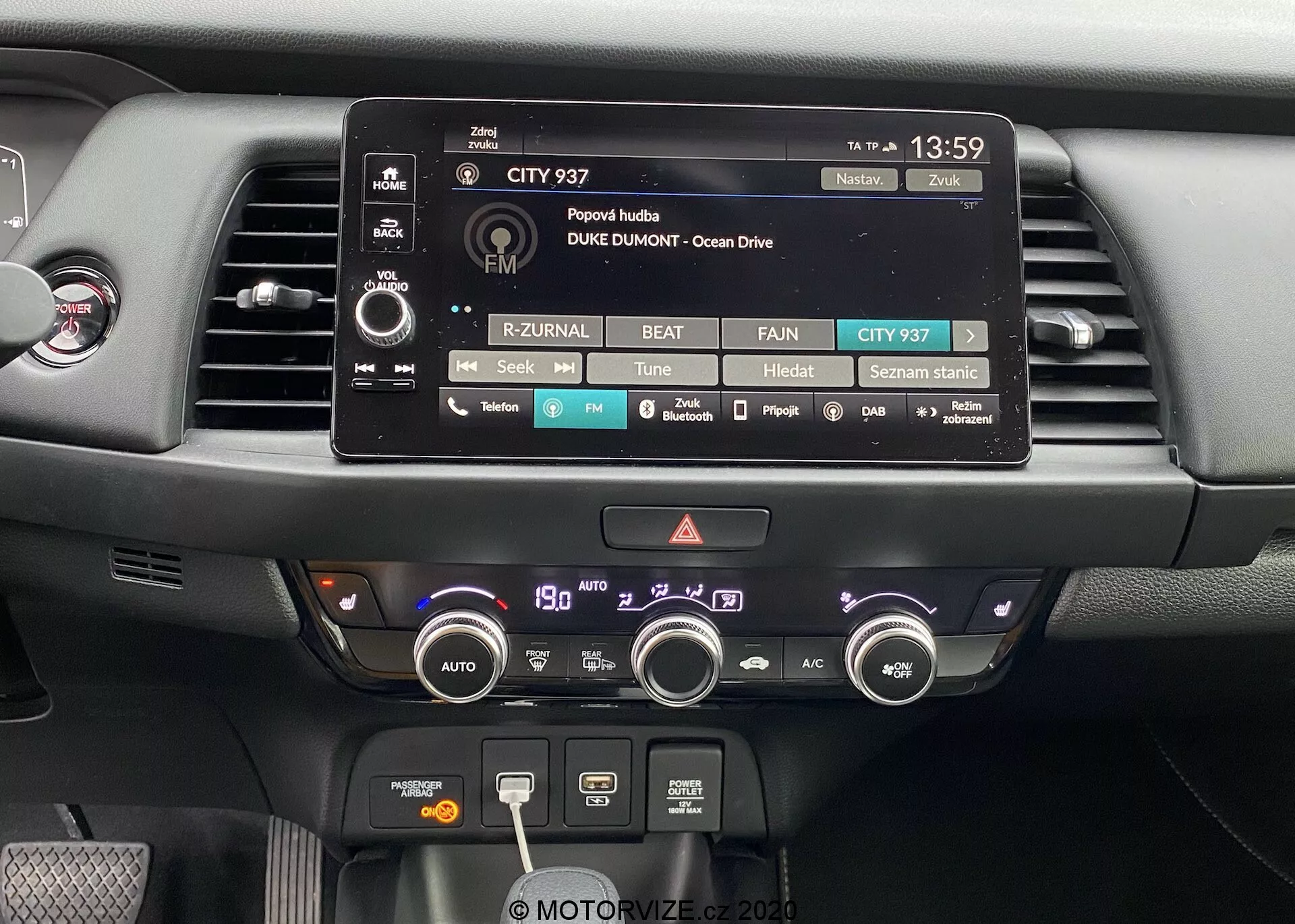 Vista ravvicinata della zona del cruscotto centrale della Honda Jazz 2020. L'immagine mette in evidenza il display touchscreen del sistema di infotainment, circondato da pulsanti e manopole fisiche per controllare l'audio, i media e le impostazioni del veicolo. Sotto ci sono due bocchette dell'aria e un sistema di controllo del clima con manopole e pulsanti per regolare la temperatura, la velocità della ventola, le funzioni di sbrinamento e la recircolazione. Il cruscotto è composto da materiali moderni, inclusi plastica opaca e lucida, progettato con l'accessibilità funzionale e la facilità d'uso in mente.