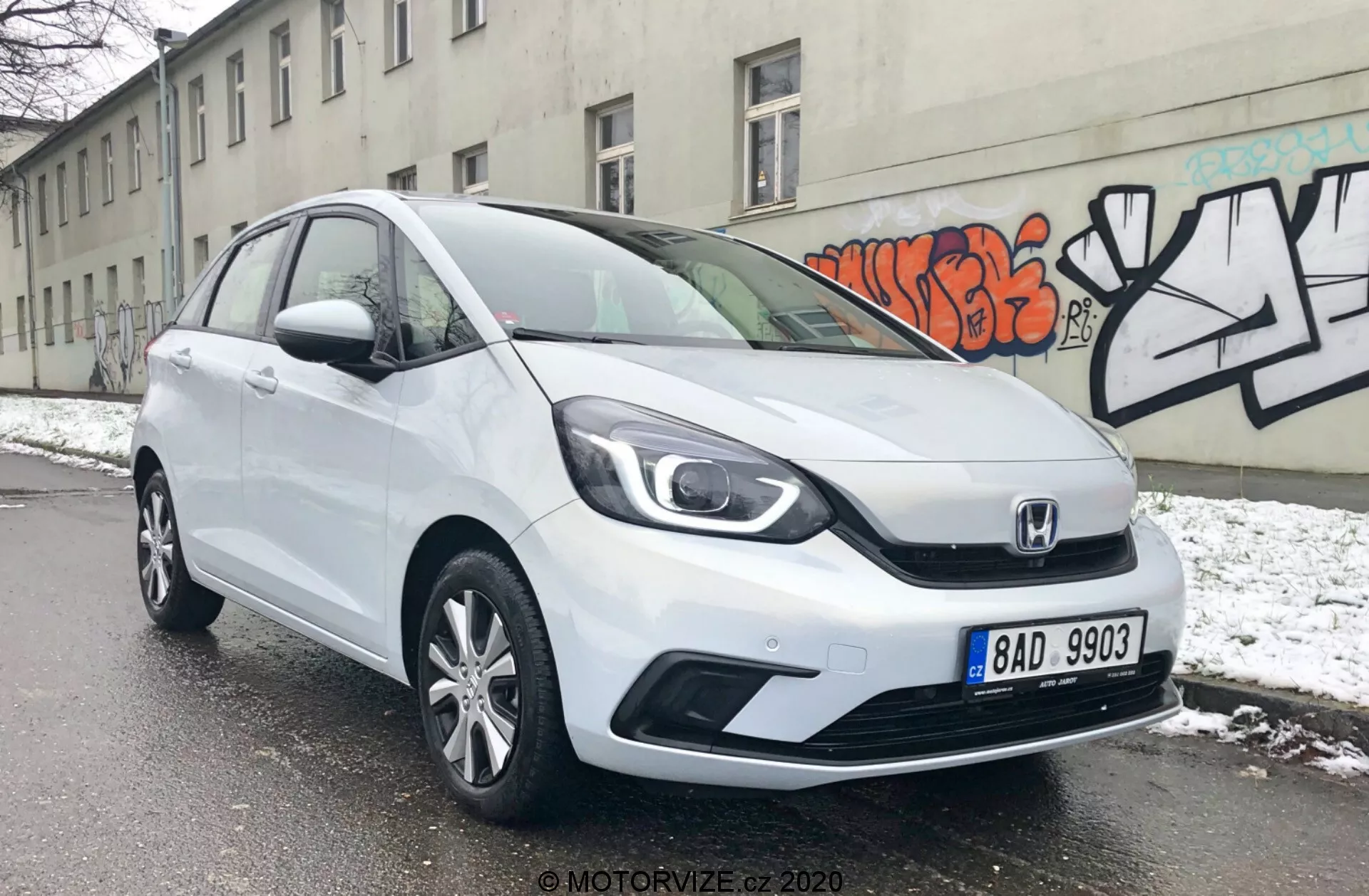 L'immagine mostra una Honda Jazz 2020 hatchback bianca vista da un angolo di tre quarti frontale, rivelando i suoi eleganti fari, la caratteristica griglia Honda con un design a due barre e le linee di contorno aerodinamiche sul cofano. Il paraurti anteriore presenta prese d'aria visibili e possibilmente fendinebbia, mentre il profilo laterale mostra ruote in lega multi-razza e pannelli porta leggermente sagomati. L'auto è parcheggiata su una strada innevata, indicando un ambiente invernale, con uno sfondo di un muro grigio decorato con graffiti, evidenziando un ambiente urbano.