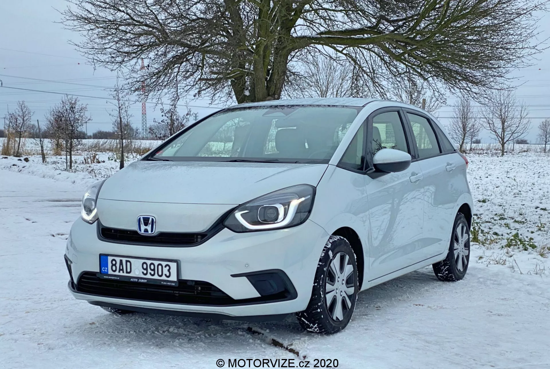 Vista angolare di tre quarti anteriore di una Honda Jazz 2020, che mette in evidenza la parte anteriore e parte del lato passeggero di questa moderna hatchback compatta. L'immagine, apparentemente scattata all'altezza degli occhi, evidenzia la griglia minimalista con il logo centrale Honda, i fari eleganti con luci diurne a LED, i cerchi in lega multirazza e gli specchietti laterali con indicatori di direzione integrati. L'auto è parcheggiata su un'area pavimentata parzialmente coperta di neve, suggerendo un'ambientazione invernale con alberi senza foglie e un cielo grigio sullo sfondo. Il veicolo appare pulito e ben mantenuto.