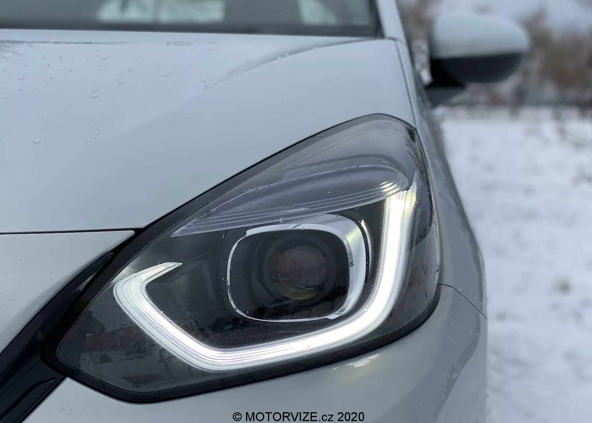 Primo piano del faro anteriore sinistro di una Honda Jazz 2020, che mostra il suo design moderno con elementi di illuminazione LED o HID. Il faro ha una luce diurna distintiva a forma di gancio, una lente del proiettore e gocce d'acqua che suggeriscono pioggia recente o lavaggio. Il design elegante è enfatizzato da linee e contorni lisci, con uno sfondo freddo o invernale, forse nevoso.
