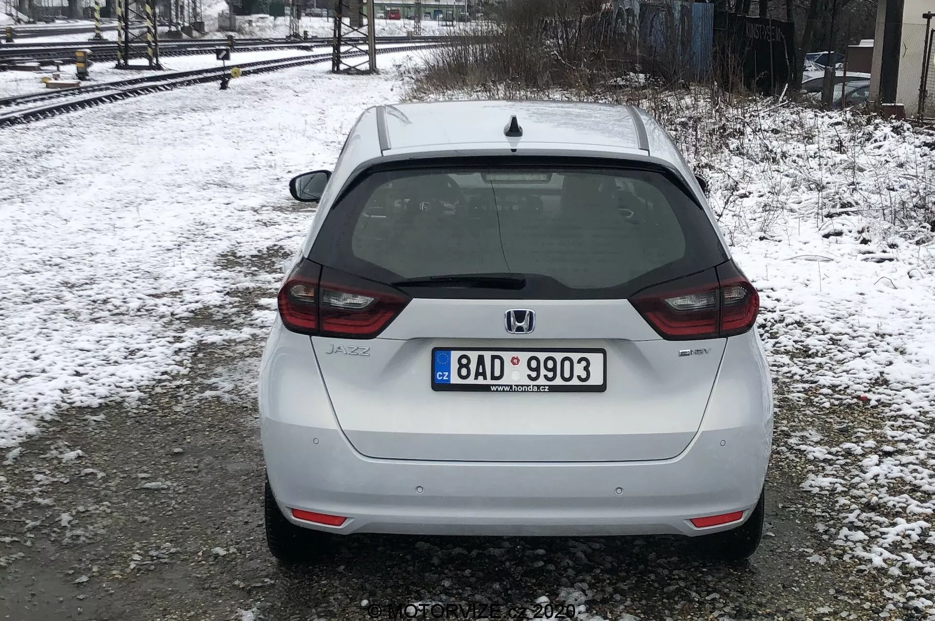 L'immagine mostra la vista posteriore di una Honda Jazz 2020 bianca, parcheggiata su un terreno innevato vicino ai binari ferroviari in condizioni di luce diffusa o nuvolosa. L'auto occupa la maggior parte dell'inquadratura, con una vista posteriore centrata che mostra la targa in stile europeo, il tergicristallo posteriore, la luce di stop montata in alto e il logo Honda. Il badge del modello 'JAZZ' è visibile nella parte inferiore sinistra del portellone, e sopra la targa è presente una telecamera posteriore. Le luci posteriori avvolgono parzialmente i lati, con riflettori nella parte inferiore del paraurti, che presenta anche una sezione integrata nera. I segni di sporcizia suggeriscono che l'auto sia stata guidata su strade bagnate o fangose. La neve e gli alberi spogli indicano che è inverno o inizio primavera.