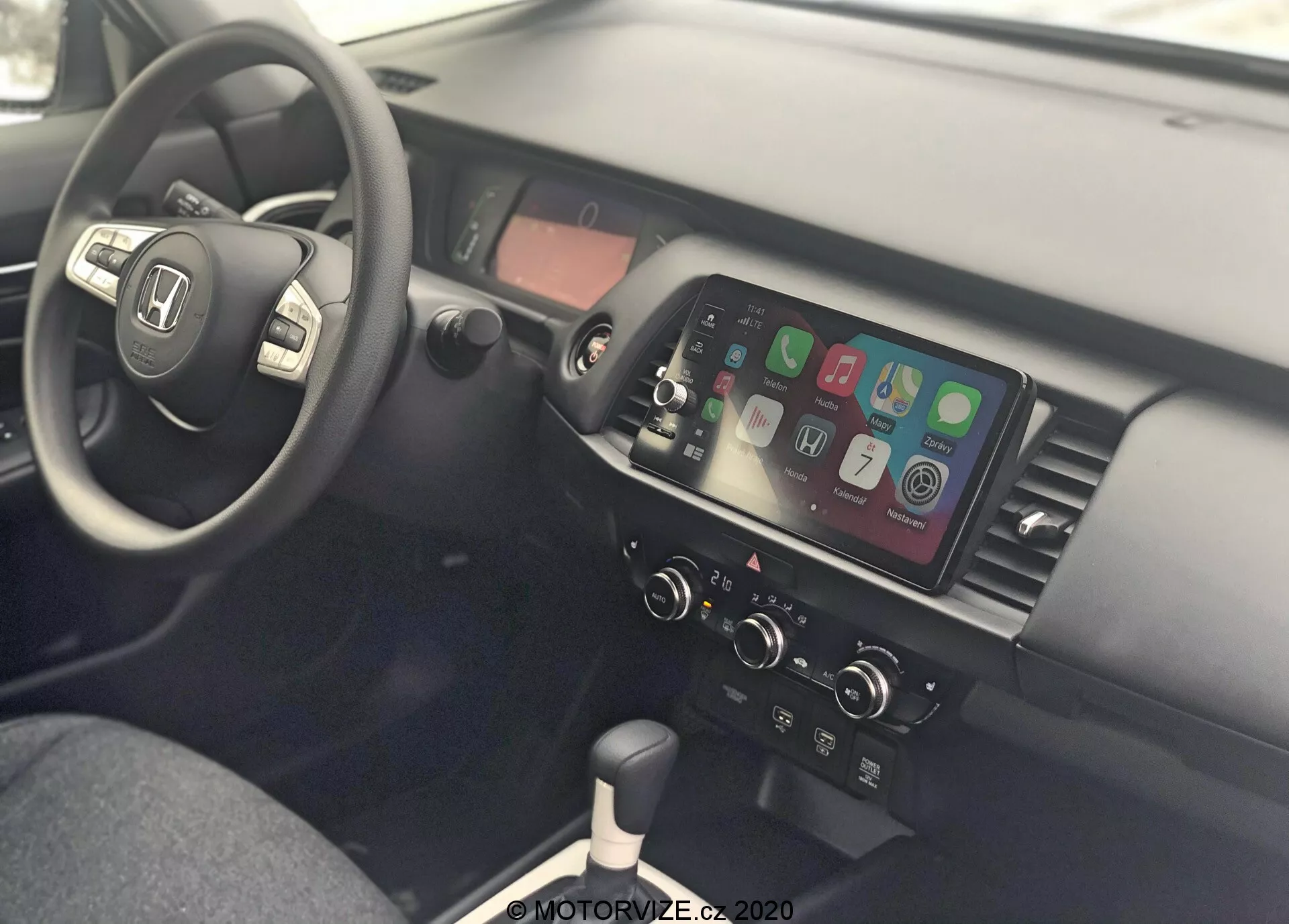 Vista interna di una Honda Jazz 2020, che mostra l'area del conducente e la console centrale dal lato passeggero. Il volante è a sinistra e presenta comandi per audio, connettività telefonica e controllo della velocità, indicando un design per la guida sul lato destro della strada. La console centrale è dotata di un display touchscreen che mostra l'integrazione dello smartphone come Apple CarPlay o Android Auto, controlli manuali del clima, leva del cambio per la trasmissione manuale, porte USB e una presa di corrente da 12V, evidenziando la connettività moderna e il design funzionale.