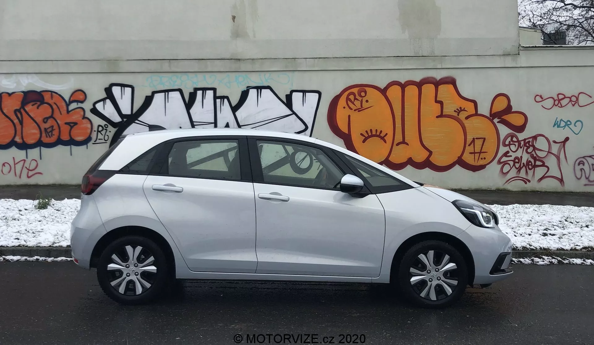 Profilo laterale di una Honda Jazz compatta bianca del 2020 parcheggiata su una strada urbana con graffiti sul muro di sfondo. La scena è durante un giorno nuvoloso con una leggera spruzzata di neve a terra, suggerendo condizioni invernali. L'immagine cattura il lato del conducente, mostrando maniglie delle porte in tinta con la carrozzeria, specchietti laterali, cerchi in lega a cinque razze e finestre standard con un montante B nero. Il veicolo appare pulito con qualche macchia umida dalla neve. È visibile il watermark 'MOTORVIZE.CZ 2020', che indica la fonte della foto.