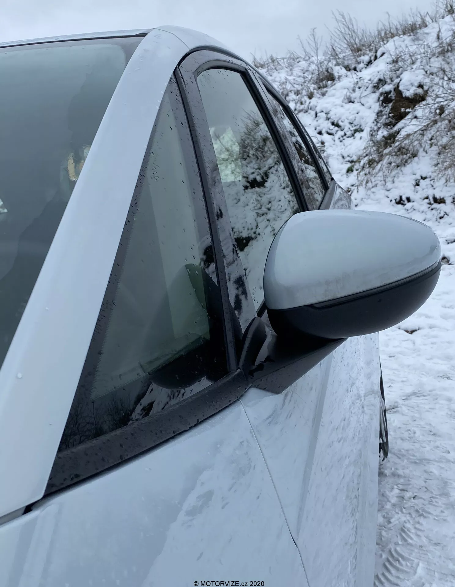 Primo piano del lato destro di una Honda Jazz 2020 che mostra l'area della portiera del guidatore. Lo specchietto laterale prominente si estende da sinistra dell'immagine al centro, suggerendo un uso recente o prossimo. La calotta dello specchietto si abbina al colore dell'auto, indicando che è una caratteristica standard. Gli elementi di design moderno includono uno specchietto aerodinamico e la carrozzeria intatta. Il finestrino è alzato con condensazione all'interno, e si possono vedere fiocchi di neve sulla portiera e sul finestrino. Il montante B visibile e parti di entrambe le portiere, anteriore e posteriore, indicano un veicolo a quattro porte. La freccia laterale non è visibile. Il fondo mostra condizioni invernali con neve a terra.