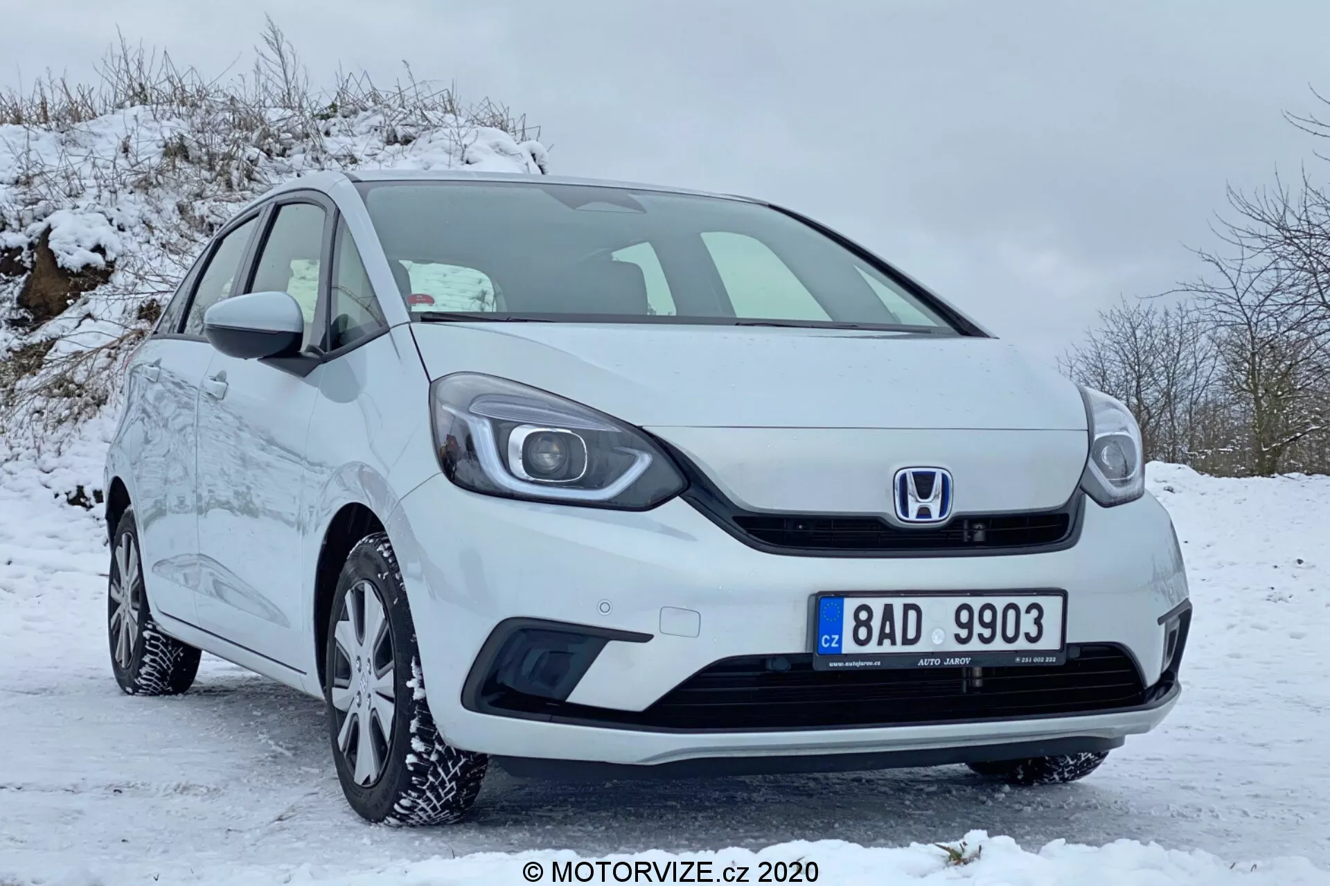 Vista frontale a tre quarti di una Honda Jazz 2020 di colore bianco, parcheggiata su un'area coperta di neve, con la griglia con l'emblema Honda, il paraurti anteriore, i fari, i cerchi in lega con pneumatici invernali, gli specchietti laterali e le maniglie delle porte, il tutto in condizioni ben mantenute e pulite.