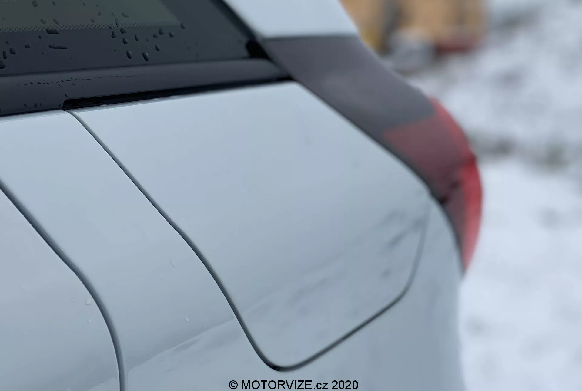 Dettaglio del Honda Jazz 2020, che mostra il quarto posteriore destro dell'auto con una vista di tre quarti laterale. Le caratteristiche includono un finestrino posteriore con linee di sbrinamento, un'antenna a pinna di squalo montata sul tetto, il montante C che accentua il design dell'auto, un parafanghi posteriore con una curva liscia, un frammento del gruppo ottico posteriore, e una finitura di vernice metallica o perlata. La poca profondità di campo mette a fuoco i dettagli del veicolo mentre lo sfondo è sfocato. La filigrana dice 'MOTORVIZE.cz, 2020.'