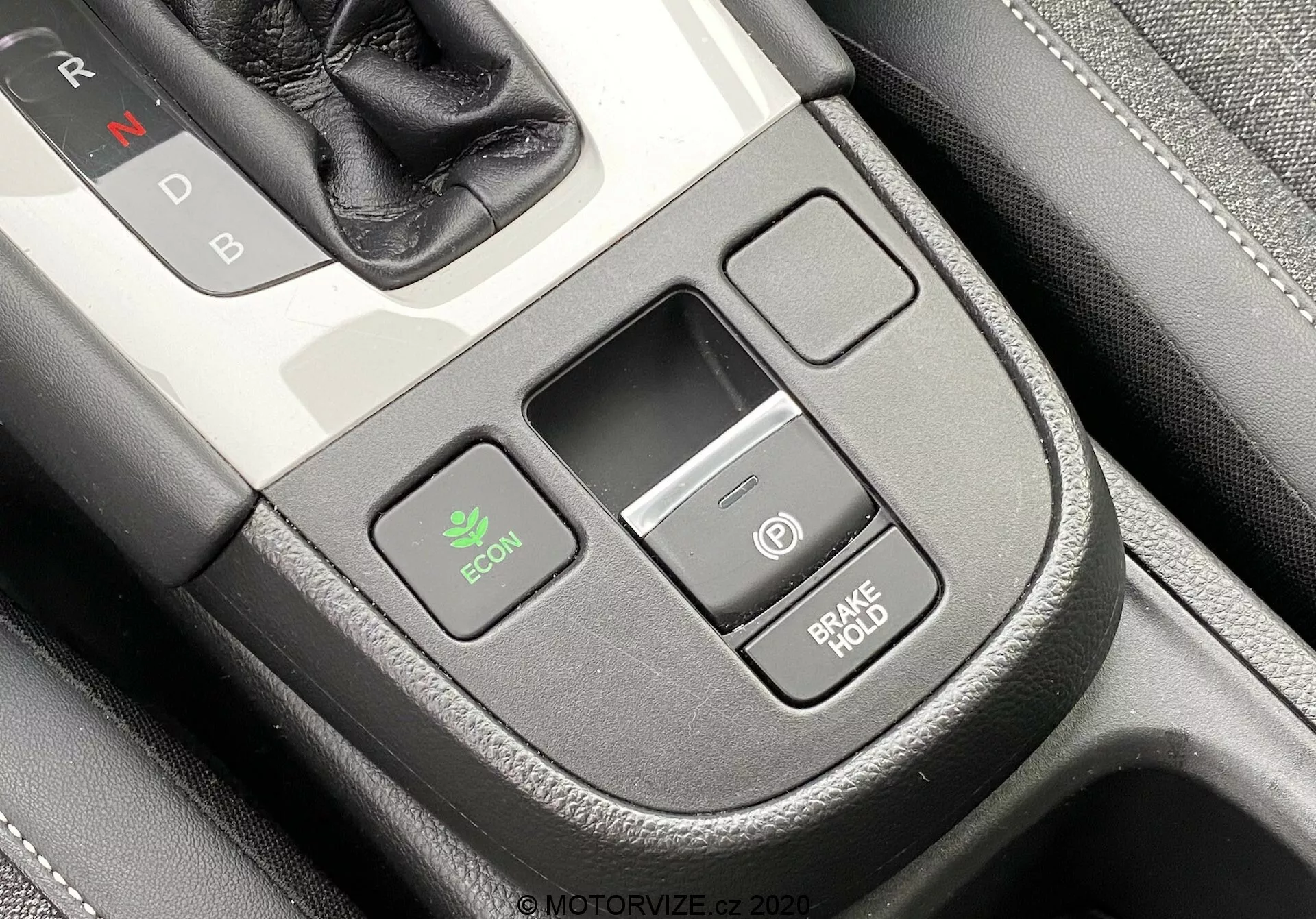 Primo piano della console centrale interna di una Honda Jazz 2020 da una leggera prospettiva dall'alto del conducente. Il selettore del cambio con 'N' di Neutrale è parzialmente visibile in alto a destra, indicando una trasmissione automatica. È evidente il pulsante ECON a sinistra per la modalità economia, una piccola fessura di archiviazione rettangolare centrale e due pulsanti sovrapposti a destra: uno con un'icona di accensione e l'altro etichettato 'BRAKE HOLD,' che previene il rollio alle fermate. La console ha una finitura premium con una texture simile al cuoio e un dettaglio di cuciture.