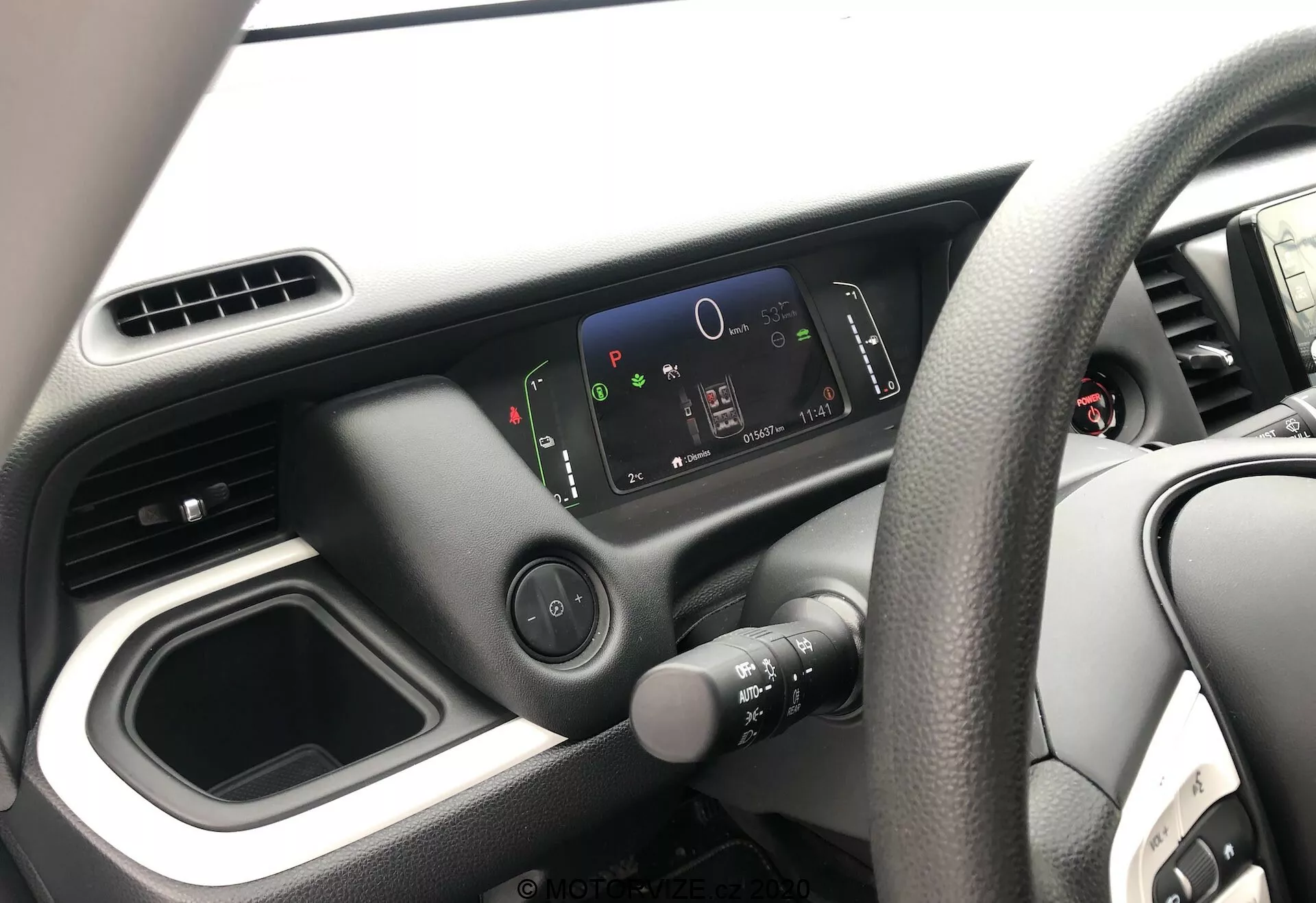 Vista interna della Honda Jazz 2020, focalizzata sul lato del conducente dalla prospettiva del passeggero. L'immagine mostra il volante a destra con un design a quattro razze, la plancia minimalista con un quadro strumenti digitale ad alta risoluzione e il semplice layout della console centrale con un vano portaoggetti vuoto e prese d'aria. È visibile solo una parte della porta del conducente.