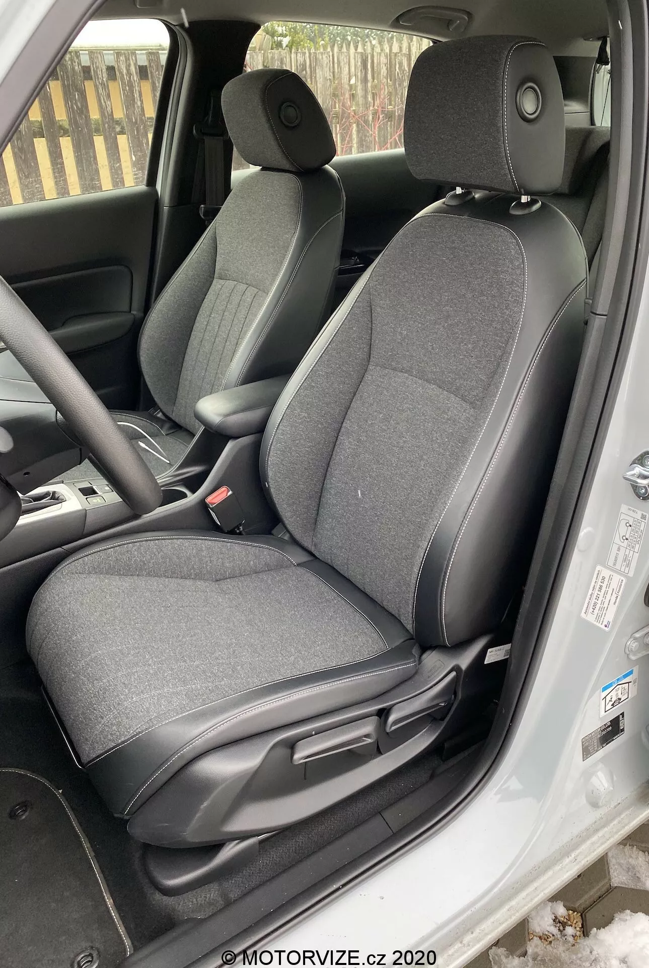 L'immagine mostra l'interno di una Honda Jazz 2020, focalizzato sull'area del sedile del conducente ripresa da una prospettiva leggermente sopra la linea degli occhi dall'esterno del veicolo. Il sedile del conducente regolabile è rivestito in tessuto scuro. Sono visibili le leve di regolazione per far scorrere e reclinare il sedile, il pannello interno della porta con controlli per i finestrini elettrici, il volante parzialmente visibile con controlli montati sul volante, una porzione del cruscotto, il tappetino del pavimento e l'area dei pedali che indicano una possibile trasmissione manuale, parte della console centrale e la cintura di sicurezza del conducente appesa al lato.