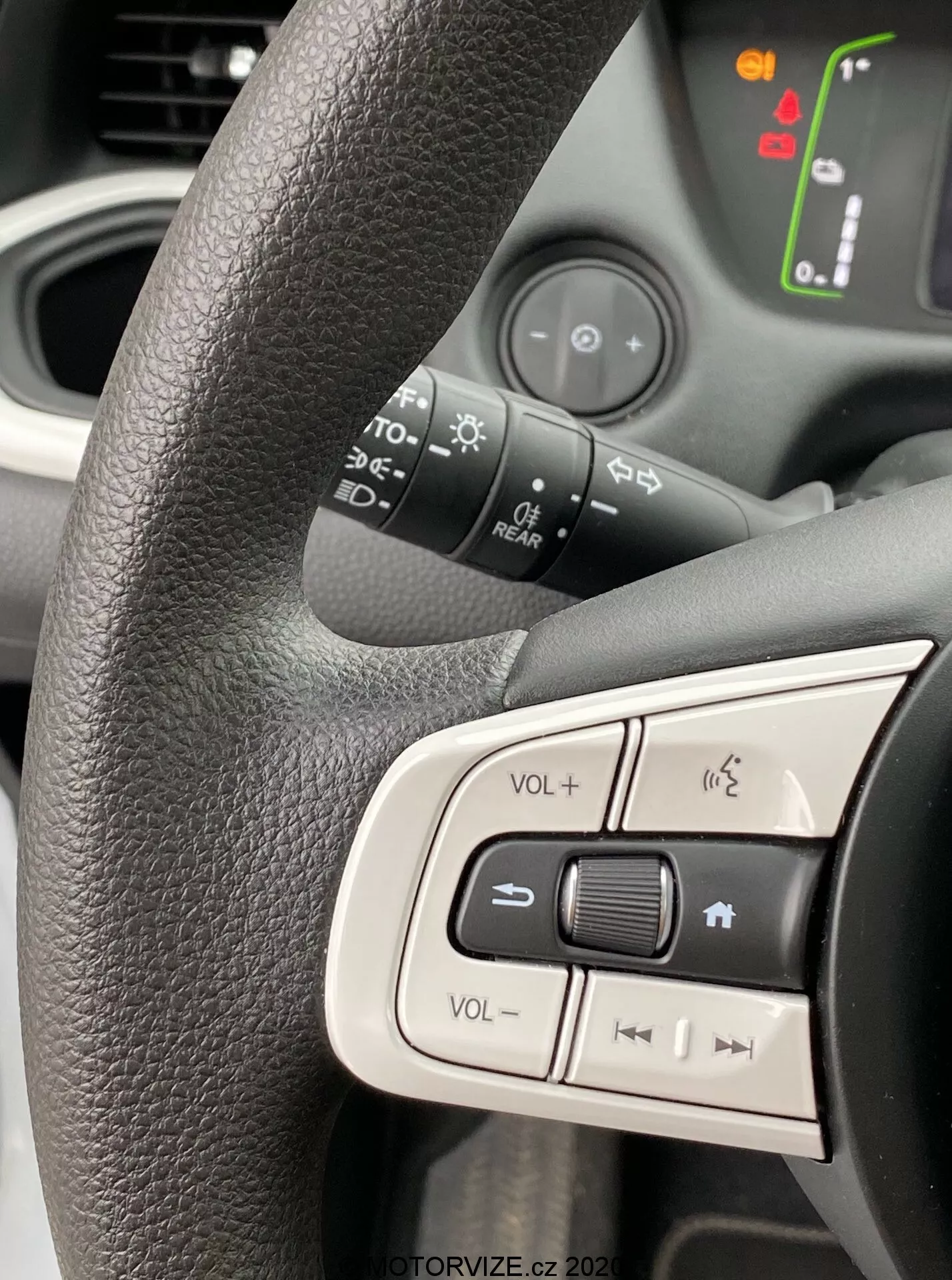 Inquadratura ravvicinata del lato sinistro del volante di una Honda Jazz 2020, focalizzata sui pulsanti di controllo integrati. Volante nero texturizzato con presa ergonomica, con pulsanti di controllo del volume, muto e navigazione multimediale. Lo sfondo leggermente sfocato rivela simboli illuminati sul cruscotto dell'auto, indicando che il veicolo è acceso. La luce naturale brillante cattura il design moderno e intuitivo, enfatizzando l'ergonomia degli interni e le caratteristiche tecnologiche.