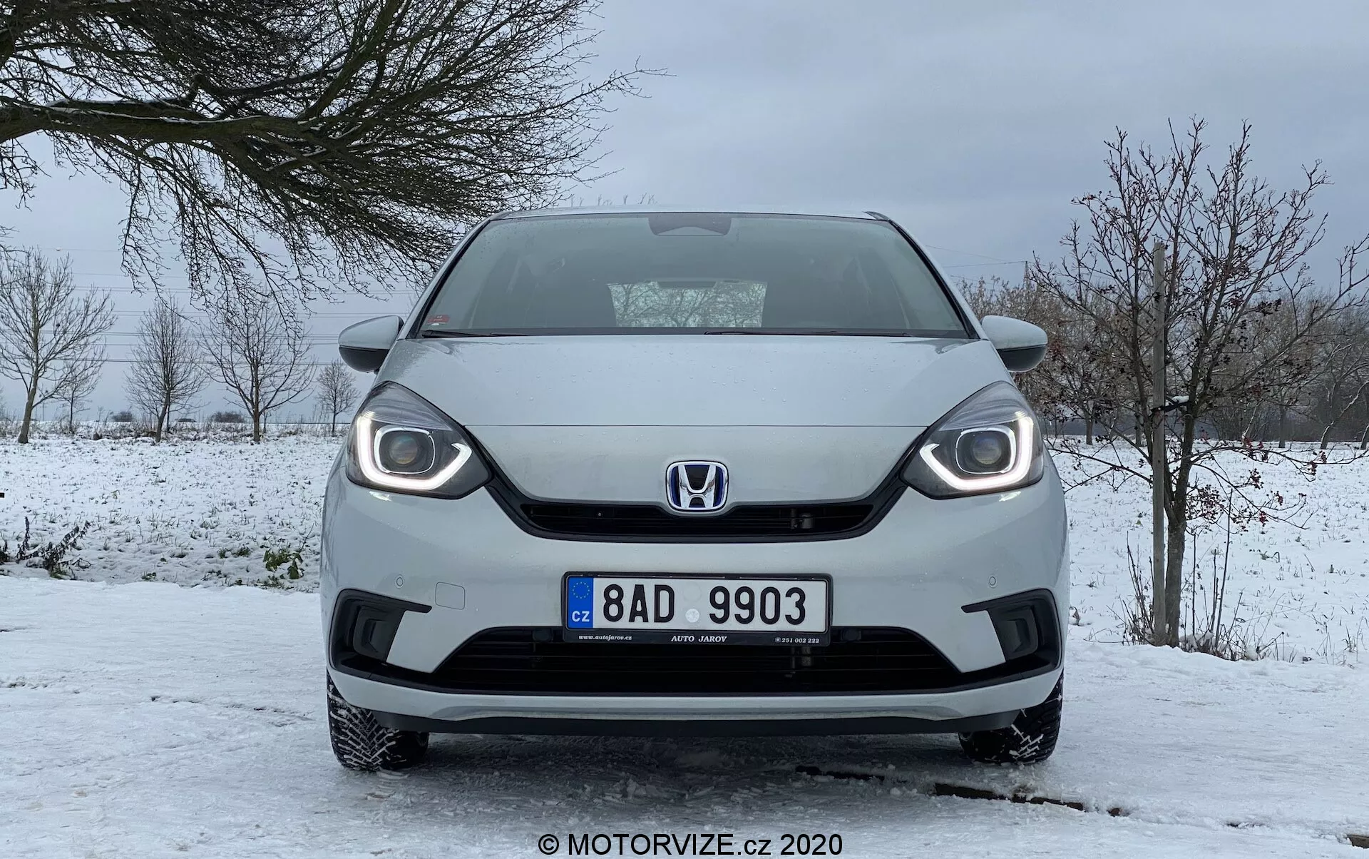 Vista frontale di una Honda Jazz 2020 in condizioni invernali, che mostra un design distintivo della griglia, fari visibili con luci diurne a LED e targa centrale, parcheggiata su una strada innevata con cielo coperto e alberi senza foglie.