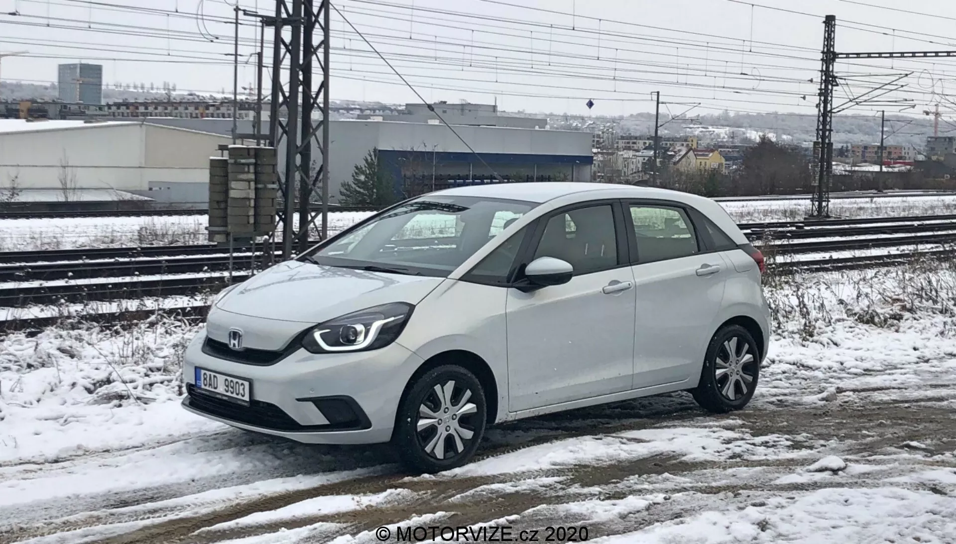 Una Honda Jazz 2020 bianca, una vettura hatchback subcompatta, catturata da una prospettiva di altezza bassa a media, che mostra sia la parte anteriore che il lato destro. La macchina è parcheggiata su una superficie nevosa e bagnata, con neve visibile sul veicolo e sul terreno, che indica condizioni climatiche fredde. Binari ferroviari e strutture industriali sono visibili sullo sfondo sotto un cielo coperto. La macchina mostra una griglia frontale principale con il logo del marchio, fari alogeni, cerchi in lega a più razze, specchietti laterali con indicatori di direzione integrati e un’antenna corta sul tetto posteriore.