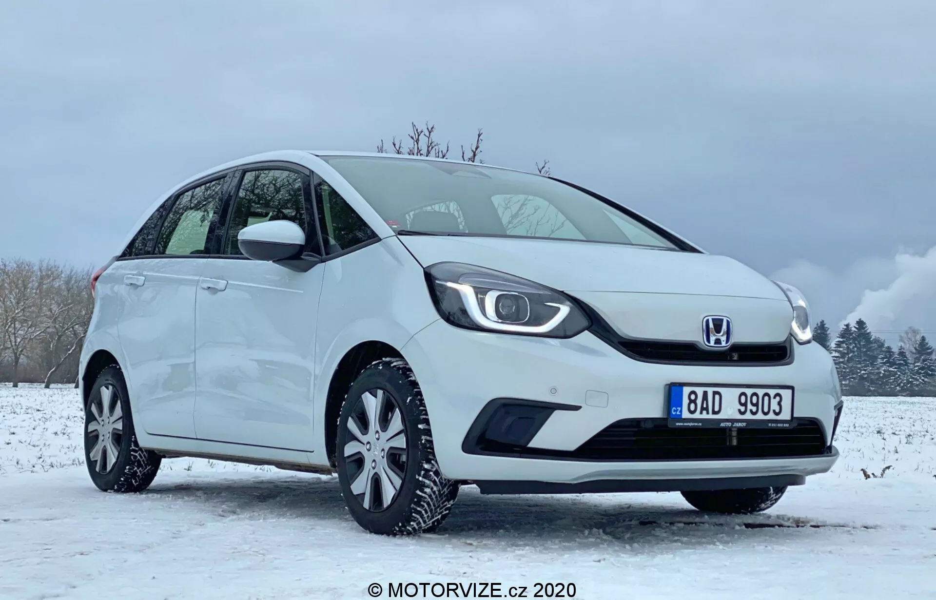 L'immagine mostra una Honda Jazz 2020 bianca, una compatta a due volumi, posizionata in un ambiente invernale con terreno innevato e cielo parzialmente nuvoloso. L'auto è vista da un'angolazione anteriore verso il lato guidatore, mettendo in risalto la griglia distintiva con il logo al centro, fari LED eleganti e paraurti anteriore liscio con alloggiamento per fendinebbia integrato. Il profilo laterale mostra una linea del tetto inclinata, cerchi in lega multi-razza e specchietti laterali e maniglie delle porte nel colore della carrozzeria. Le condizioni perfette del veicolo suggeriscono che potrebbe essere un'immagine promozionale o di recensione catturata nel 2020 da MOTORVIZE.cz.