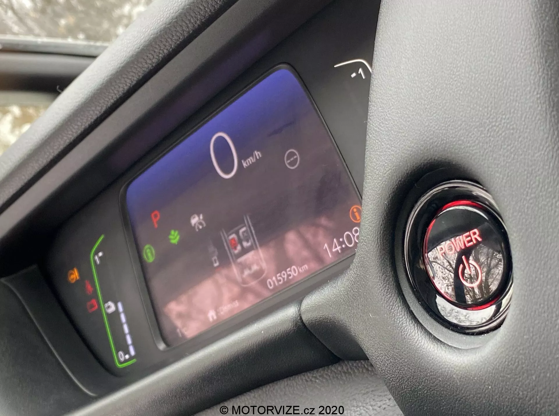 Vista ravvicinata del quadro strumenti digitale avanzato del conducente e del pulsante di avvio/arresto del motore all'interno del moderno abitacolo della Honda Jazz 2020, scattata dalla prospettiva del sedile del conducente e focalizzata sugli elementi del cruscotto. Lo schermo digitale del quadro strumenti mostra un indicatore di potenza con icone ecologiche, un tachimetro centrale a 0 km/h, diverse spie di avvertimento, un orologio e un contachilometri che segna 015902 km. Accanto ad esso si trova il pulsante di avvio del motore con anello rosso per l'accensione senza chiave.