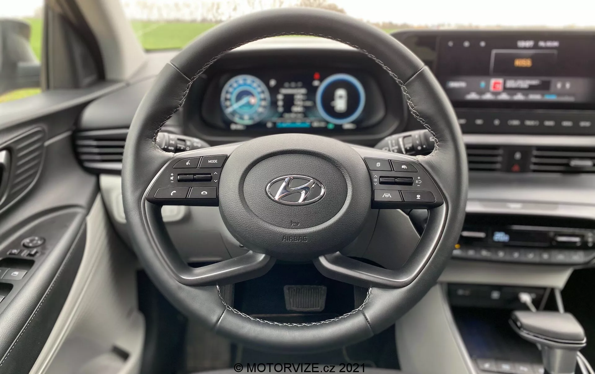 Vista interna della Hyundai i20 2020 dal punto di vista del conducente, che mostra un volante moderno a tre razze con il logo Hyundai, pulsanti multifunzione e un cruscotto completamente digitale che visualizza tachimetro, contagiri e schermo delle informazioni del conducente. Il cruscotto presenta un display centrale per l'infotainment con opzioni di menu, che indica una sofisticata integrazione tecnologica e un'interfaccia user-friendly. Il design degli interni è moderno, con linee pulite e materiali di qualità visibili nella console centrale e nella porta lato passeggero.