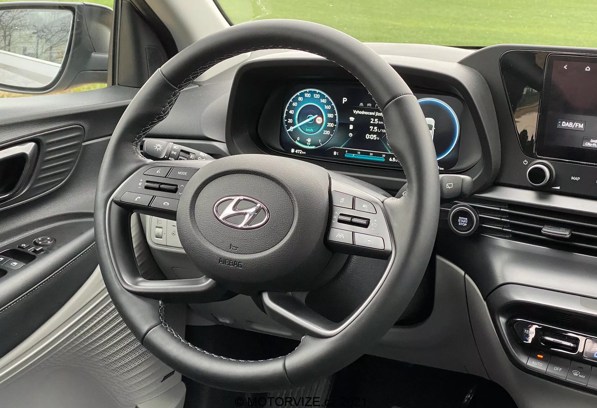 Vista interna della Hyundai i20 2020 dal sedile del conducente, che mostra il volante a tre razze con vari pulsanti di controllo, un logo del produttore ben visibile, un cruscotto digitale con schema di colori blu e bianco, tachimetro impostato su 0 km/h, selezione del cambio su Park, parte della console centrale con schermo di infotainment che visualizza la mappa del navigatore, pulsanti di controllo per clima e impostazioni del veicolo, cruscotto soft-touch con accenti metallici intorno alle bocchette dell'aria e al volante, e vista parziale della bocchetta dell'aria lato passeggero.