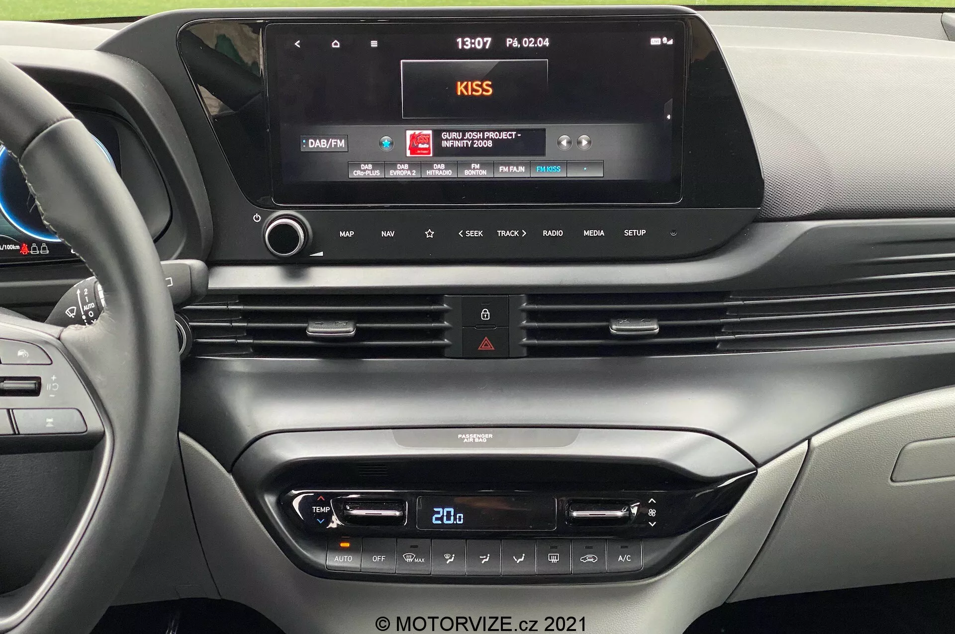 Fotografia del cruscotto interno di un'auto moderna dalla prospettiva del posto di guida, evidenziando la console centrale con un grande display di infotainment, prese d'aria integrate, pulsante delle luci di emergenza e un elegante pannello di controllo del clima con display digitale della temperatura e pulsanti sensibili al tocco. Il design del cruscotto presenta uno stile sofisticato con materiali di alta qualità e accenti metallici. Il bordo del volante con pulsanti di controllo integrati è parzialmente visibile a sinistra.