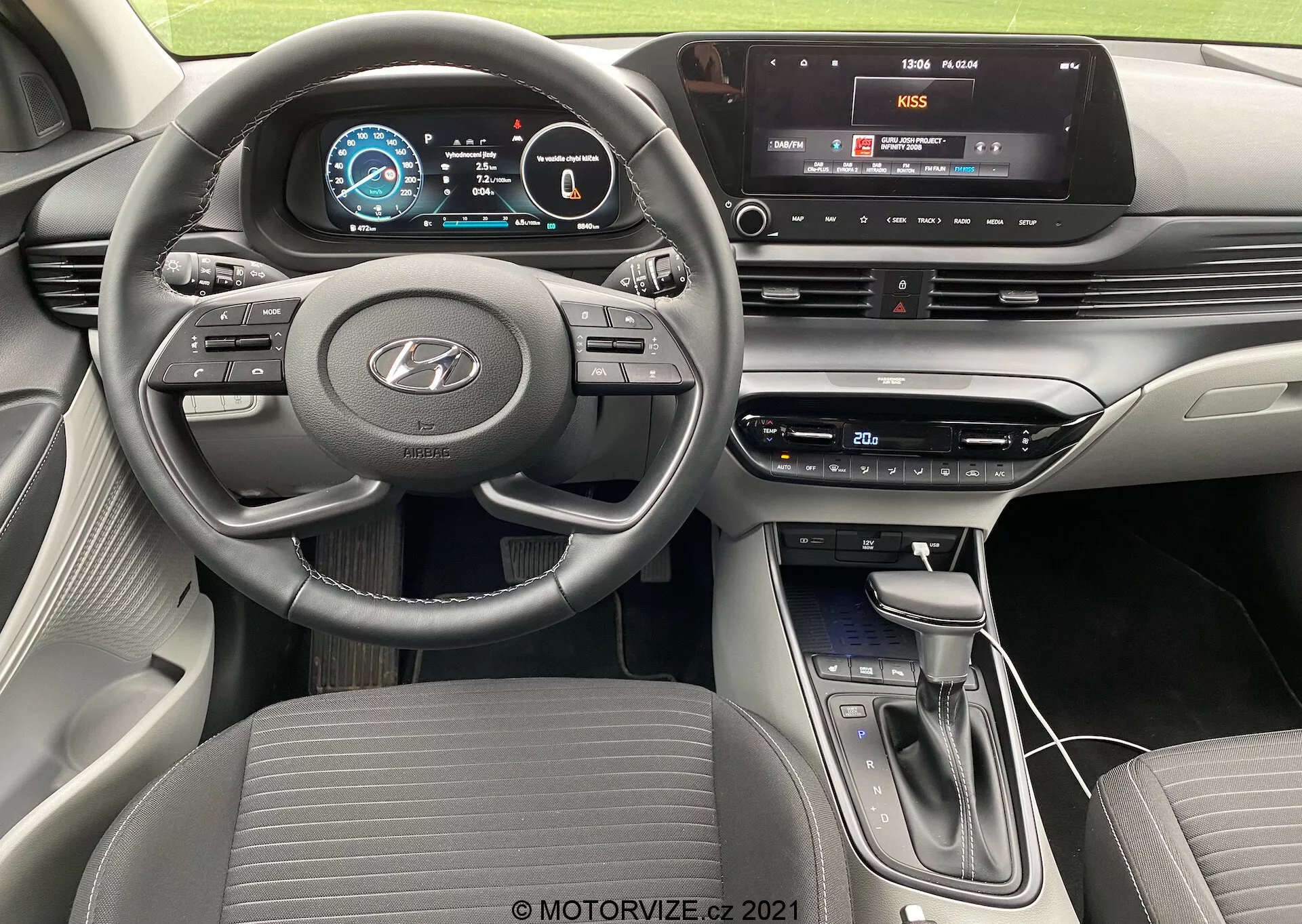 Vista dell'interno della Hyundai i20 2020 dalla prospettiva del conducente, che mostra il volante a tre razze con fondo piatto e comandi integrati per audio, controllo della velocità e comunicazione hands-free, un cruscotto completamente digitale con grafica a colori e una console centrale dotata di sistema di infotainment touchscreen con connettività multimediale, pulsanti e manopole fisici per il controllo del clima, leva del cambio moderna e design contemporaneo del cruscotto a strati con schema a due toni.