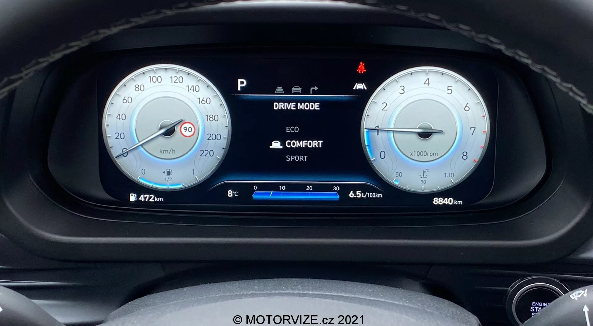Vista ravvicinata del quadro strumenti della Hyundai i20 2020 dalla prospettiva del conducente, con due indicatori analogici rotondi principali e un display digitale centrale. L'indicatore a sinistra mostra un tachimetro contrassegnato fino a 260 km/h, mentre quello a destra è un tachimetro con una linea rossa a 6500 giri/min. Il display digitale centrale mostra opzioni di modalità di guida, temperatura esterna, posizione della marcia ed efficienza del carburante. L'odometro segna 8840 km. Sono visibili varie spie, inclusa una per la funzione di start/stop automatico. L'immagine è nitida e ben illuminata, con un leggero ammorbidimento intorno ai bordi degli indicatori. Include la filigrana 'MOTORVIZE.cz 2021'.