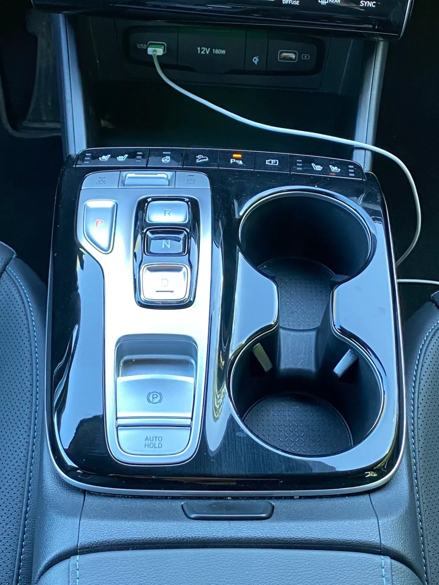 L'immagine mostra la console centrale di una Hyundai Tucson 2020, con un selettore di marcia elettronico impostato su 'N' incorniciato da una finitura nera lucida. A sinistra ci sono i comandi, tra cui un pulsante di accensione, selettori delle modalità di guida e la funzione 'AUTO HOLD'. A destra, ci sono doppi portabicchieri con un cavo di ricarica bianco in uno di essi. Nella parte superiore si intravede parte di uno schermo di infotainment con indicatori di porta USB, evidenziando la tecnologia e il lusso del veicolo.